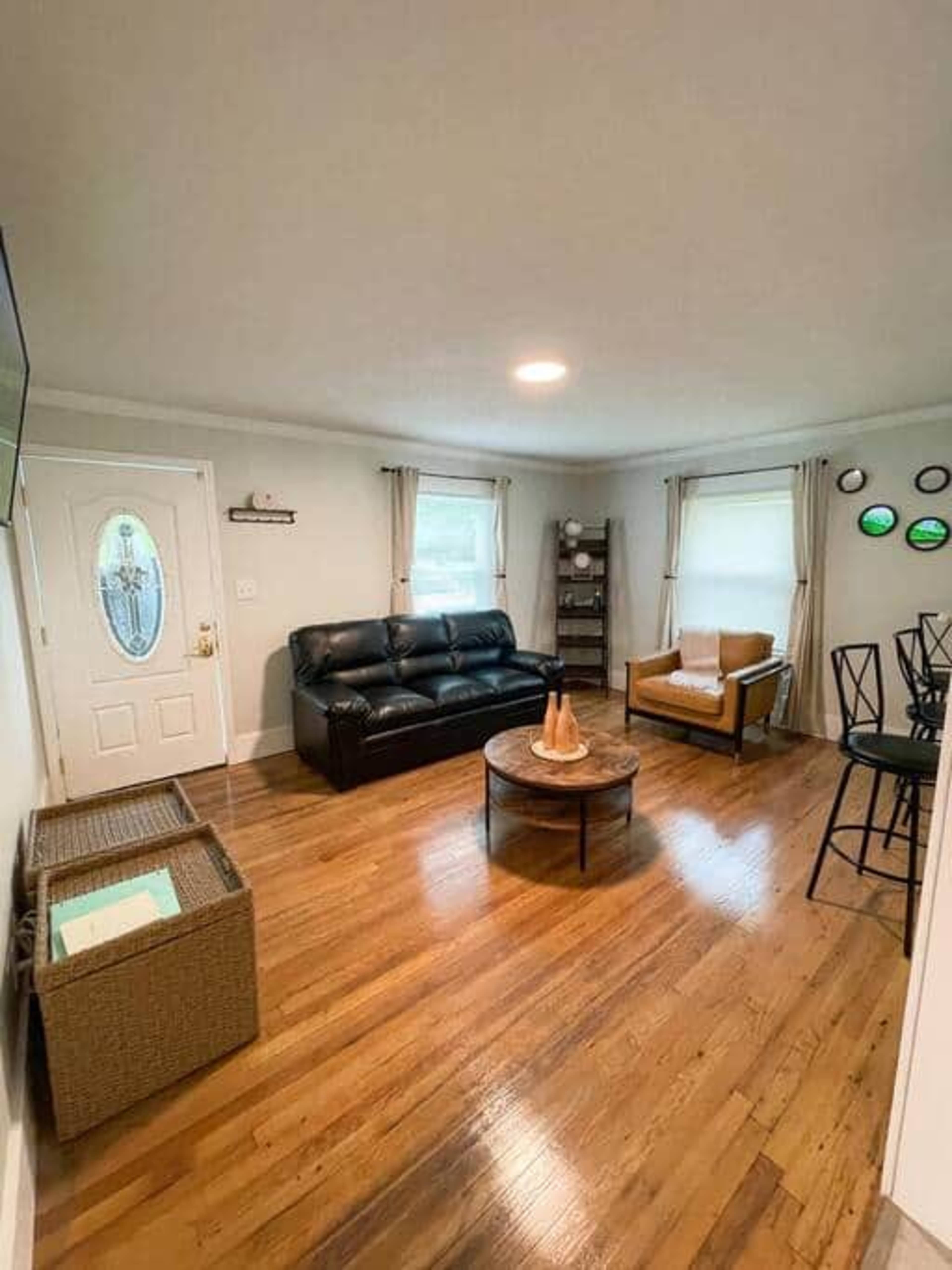 A living room features a black sofa, a brown armchair, a circular wooden coffee table, and a door leading outside.