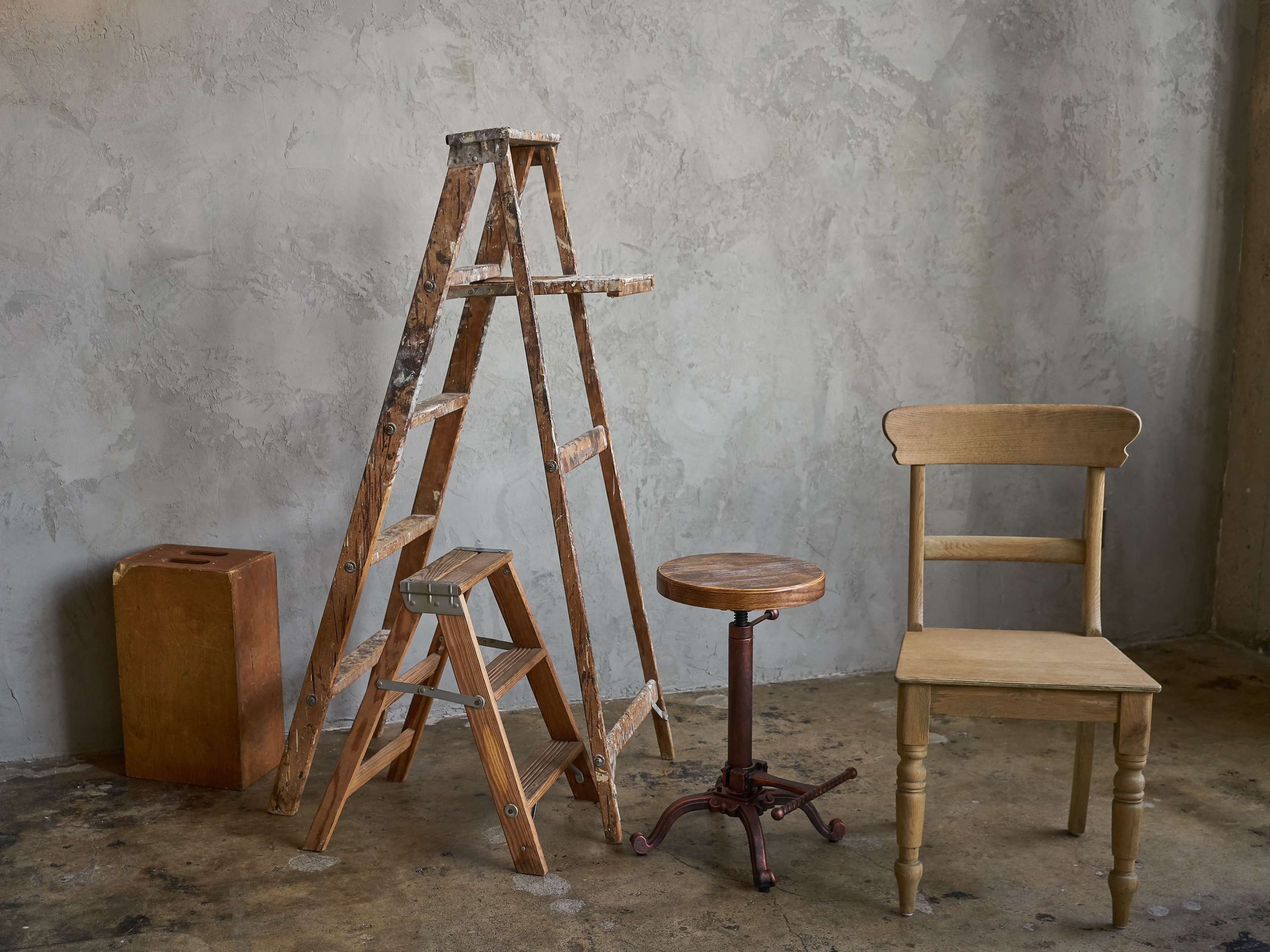 A wooden ladder, a round wooden stool, a simple wooden chair, and a small wooden box arranged against a textured gray wall.