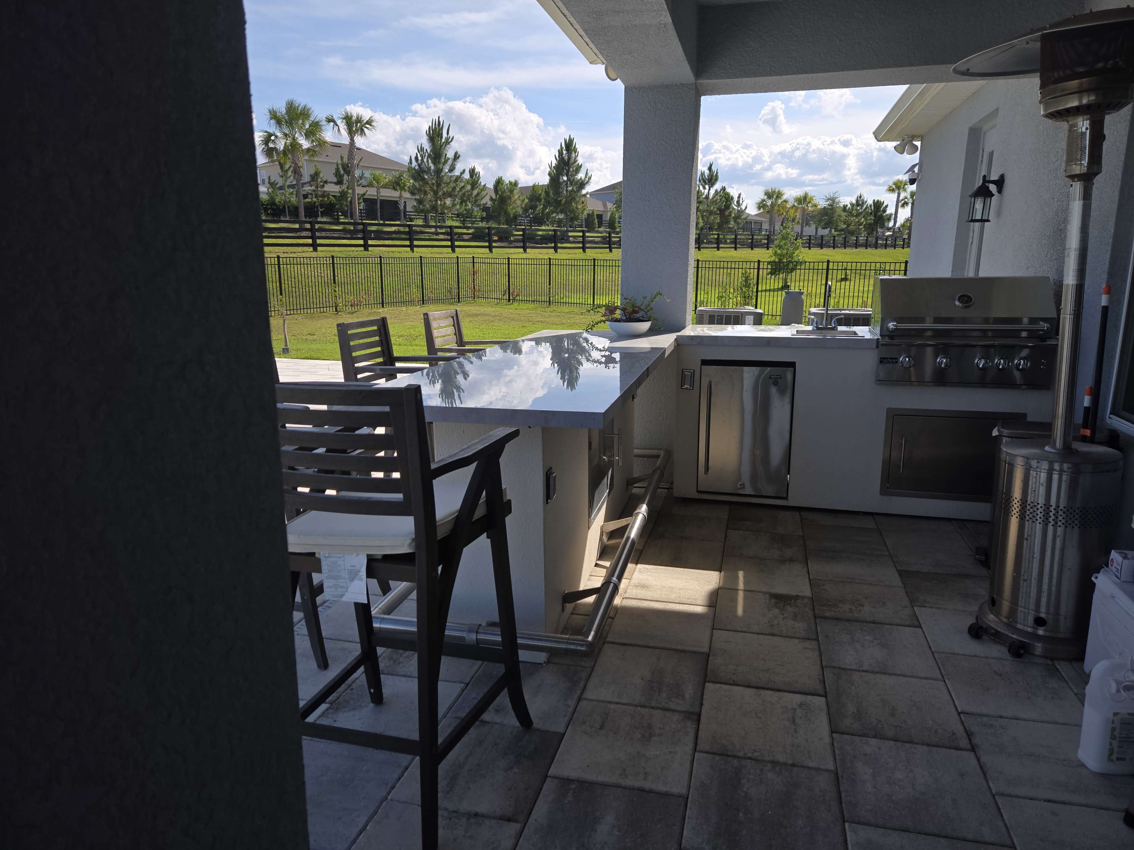 The image shows an outdoor kitchen area with a stainless steel grill, a bar-style countertop with stools, and a view of a green lawn and a fenced property in the background.