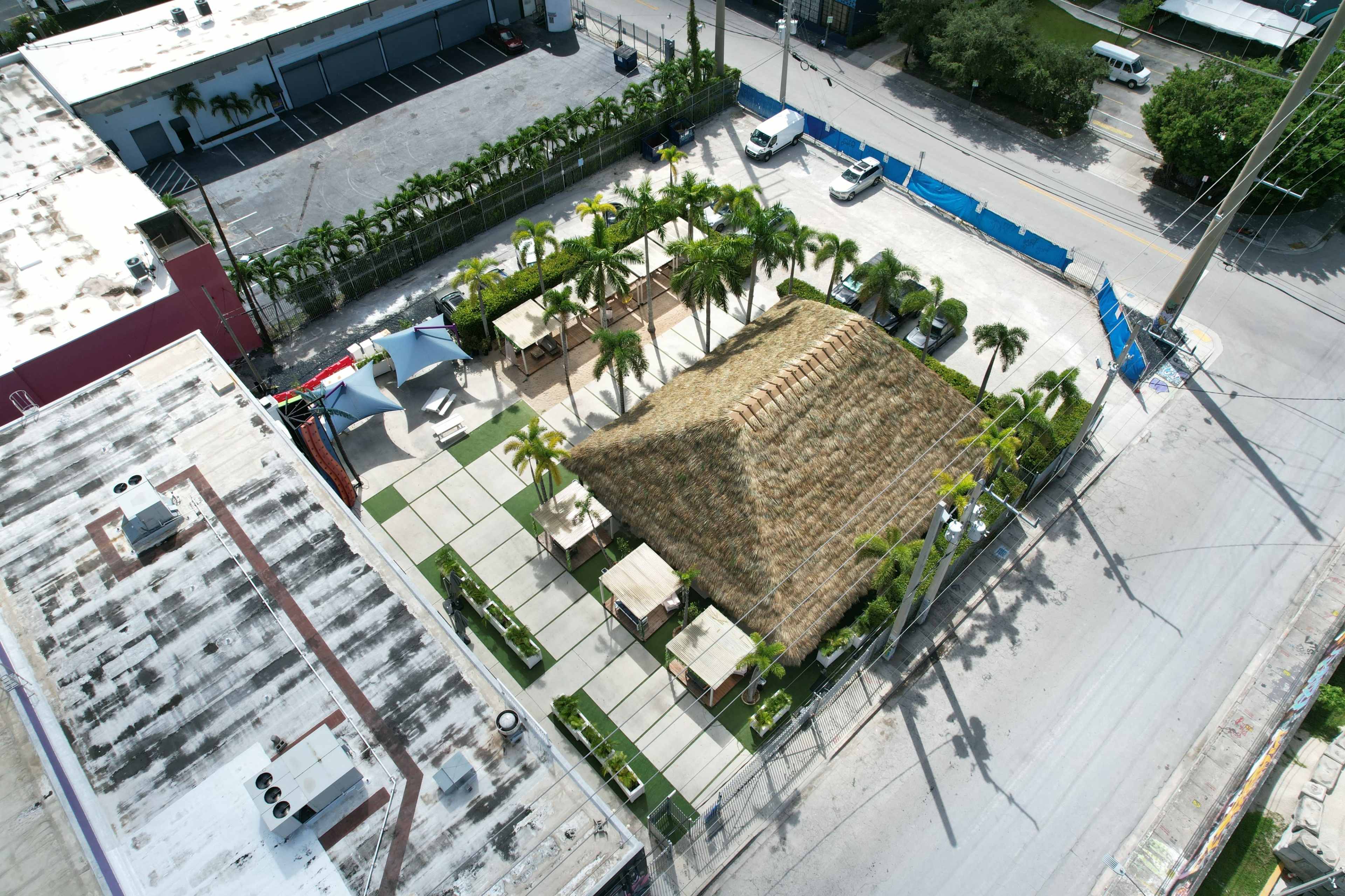 The image shows an aerial view of a tropical-themed outdoor space with a thatched-roof structure surrounded by palm trees and paved walkways.