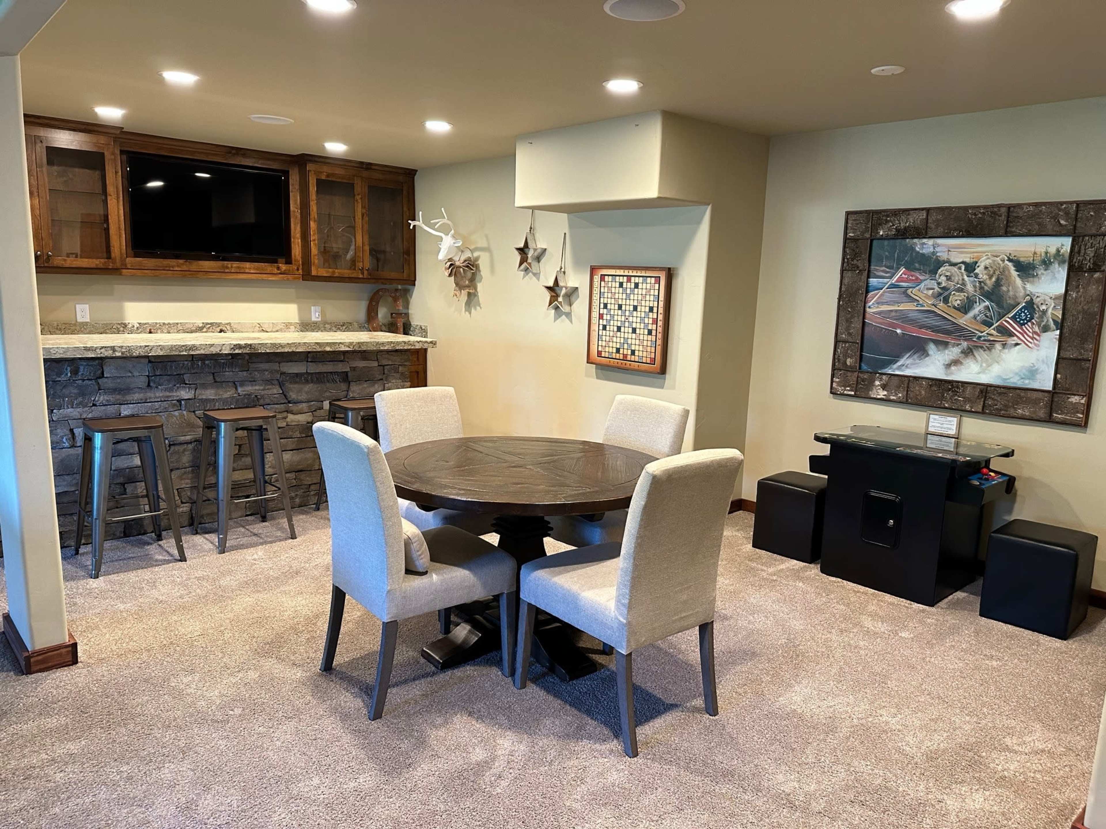 A cozy basement room featuring a circular dining table with four upholstered chairs, a stone bar area, and a decorative wall with a framed picture and various wall accents.