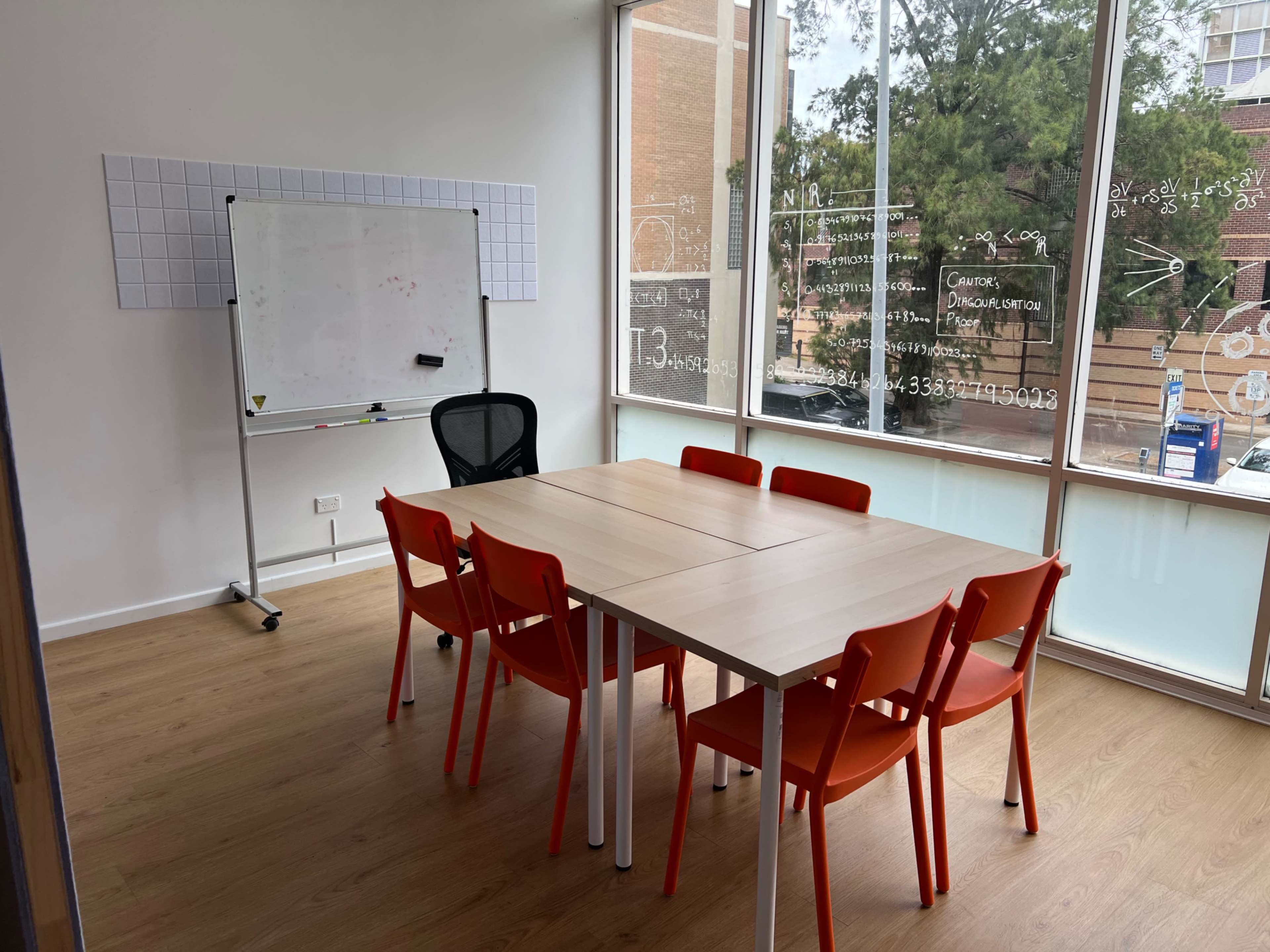 The image shows a well-lit meeting room featuring a large table surrounded by six orange chairs and a whiteboard adjacent to a window with views of greenery outside.