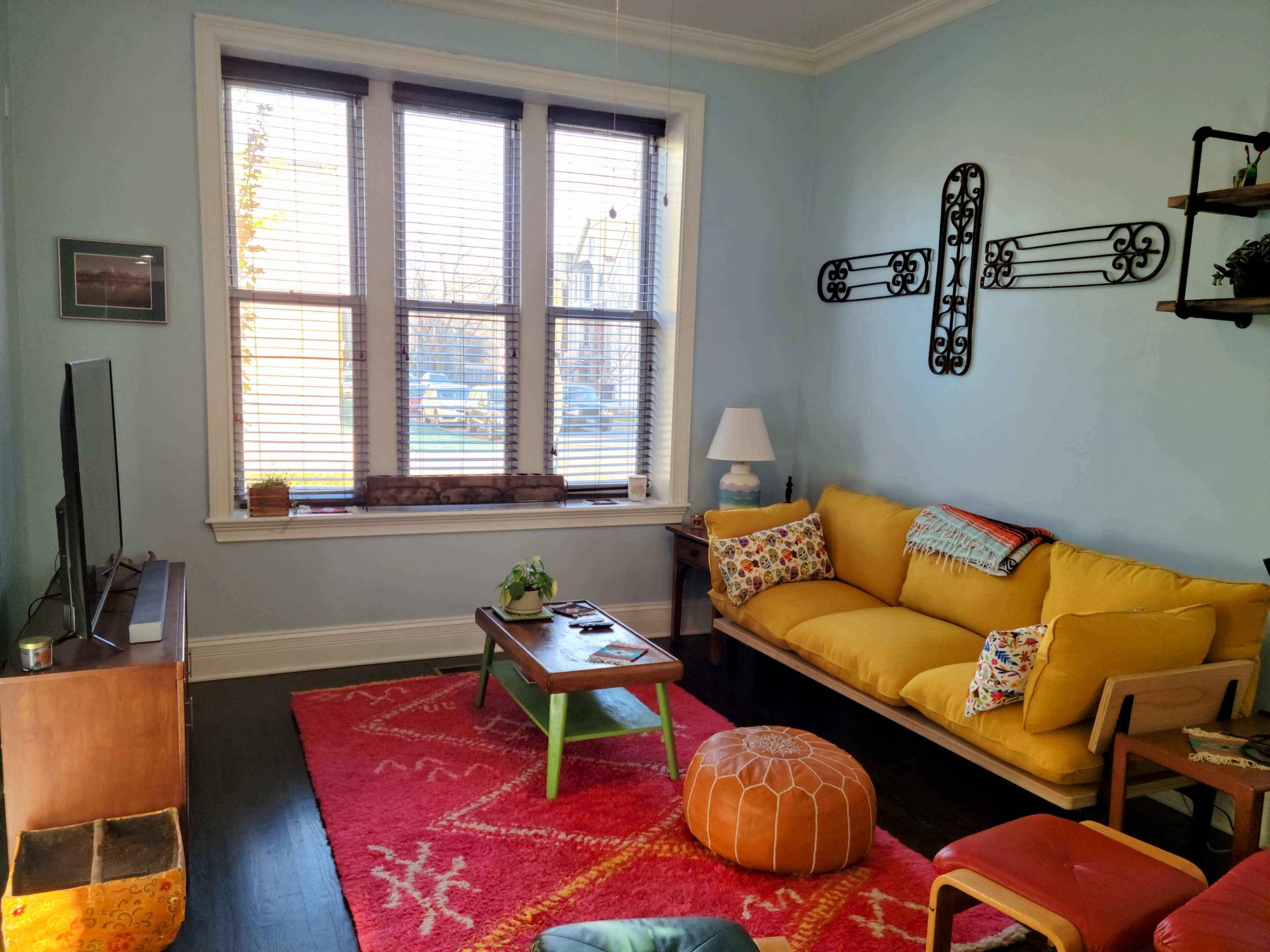 The living room features a yellow sofa, a wooden coffee table, a television, and two large windows dressed with blinds, showcasing a vibrant rug and decorative items.