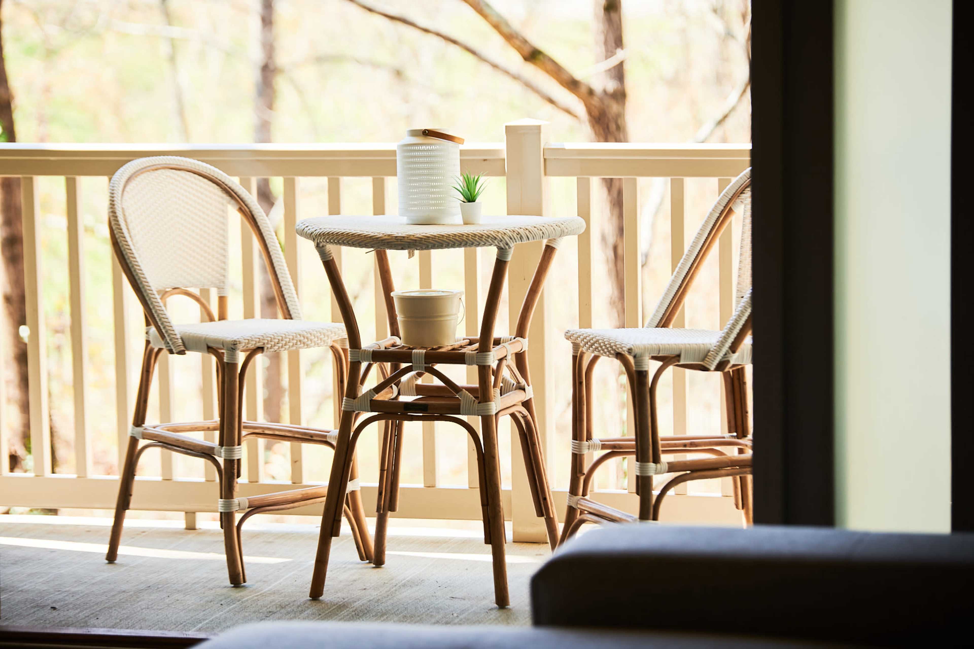 A small outdoor balcony features a round table and two chairs made of woven material, surrounded by trees in the background.