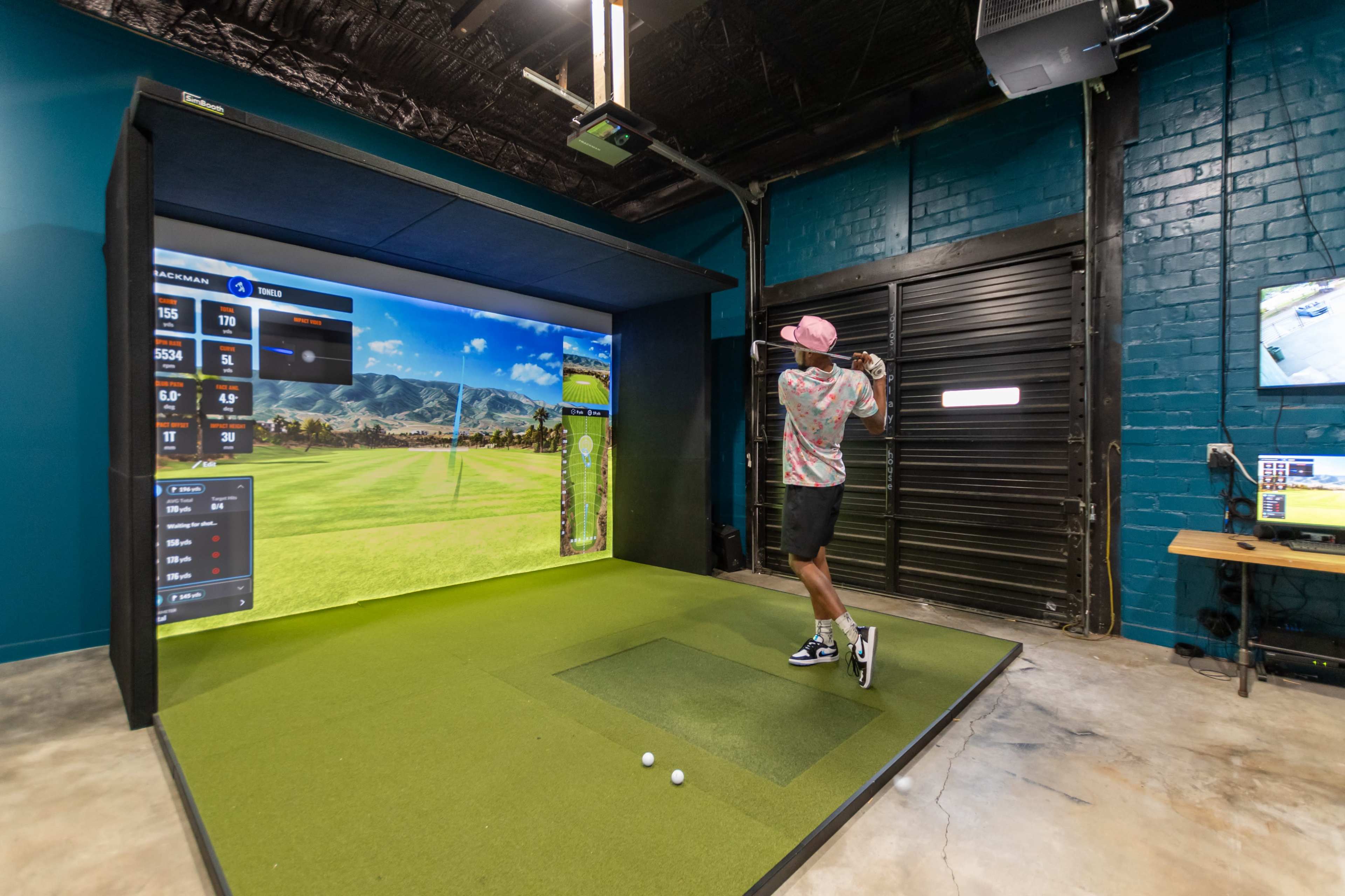 A golfer swings a club in a golf simulator room with a large screen displaying a virtual course.