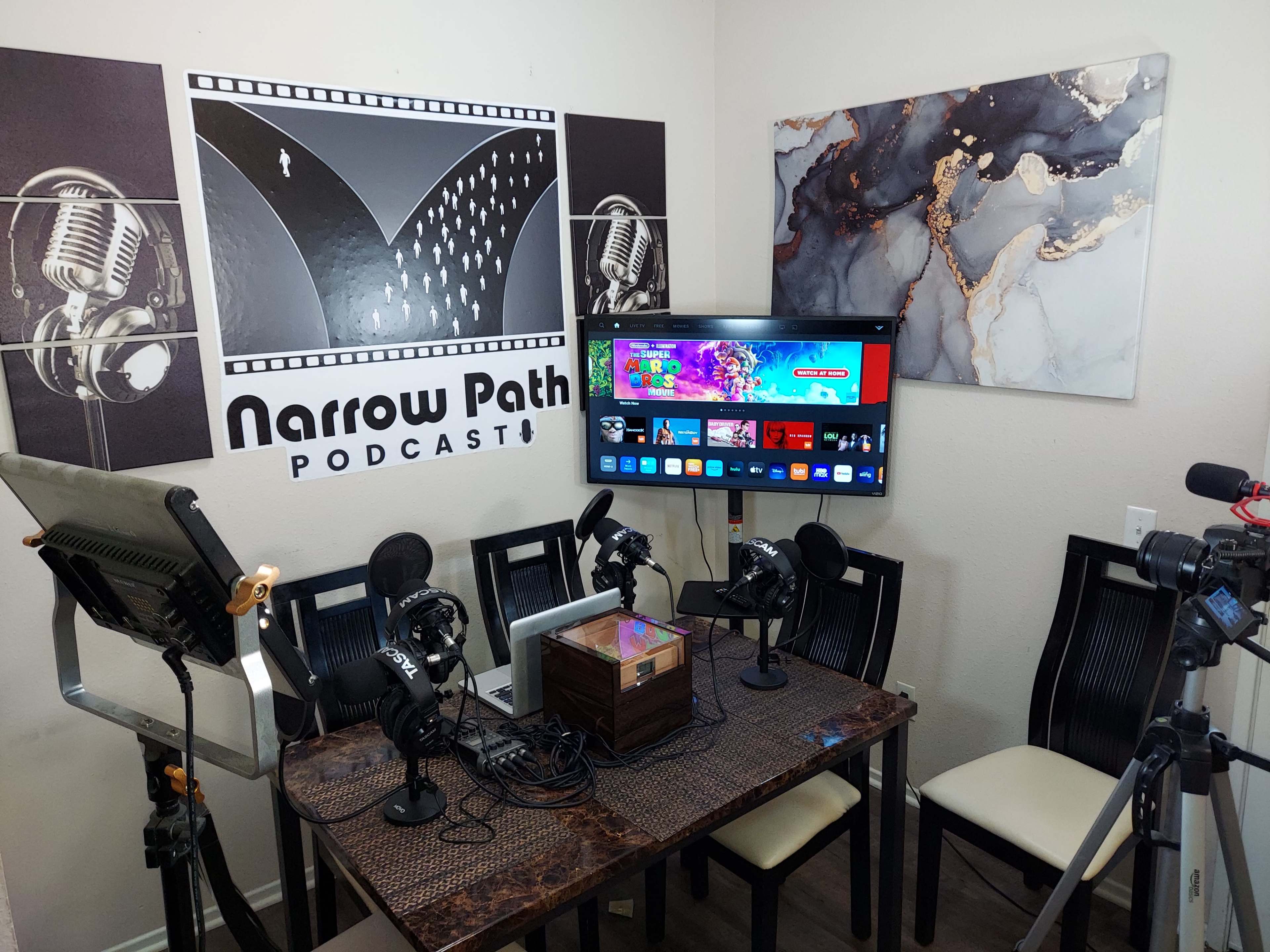 The image shows a podcasting setup featuring a table with microphones, a television displaying content, and a backdrop with the text "Narrow Path PODCAST."