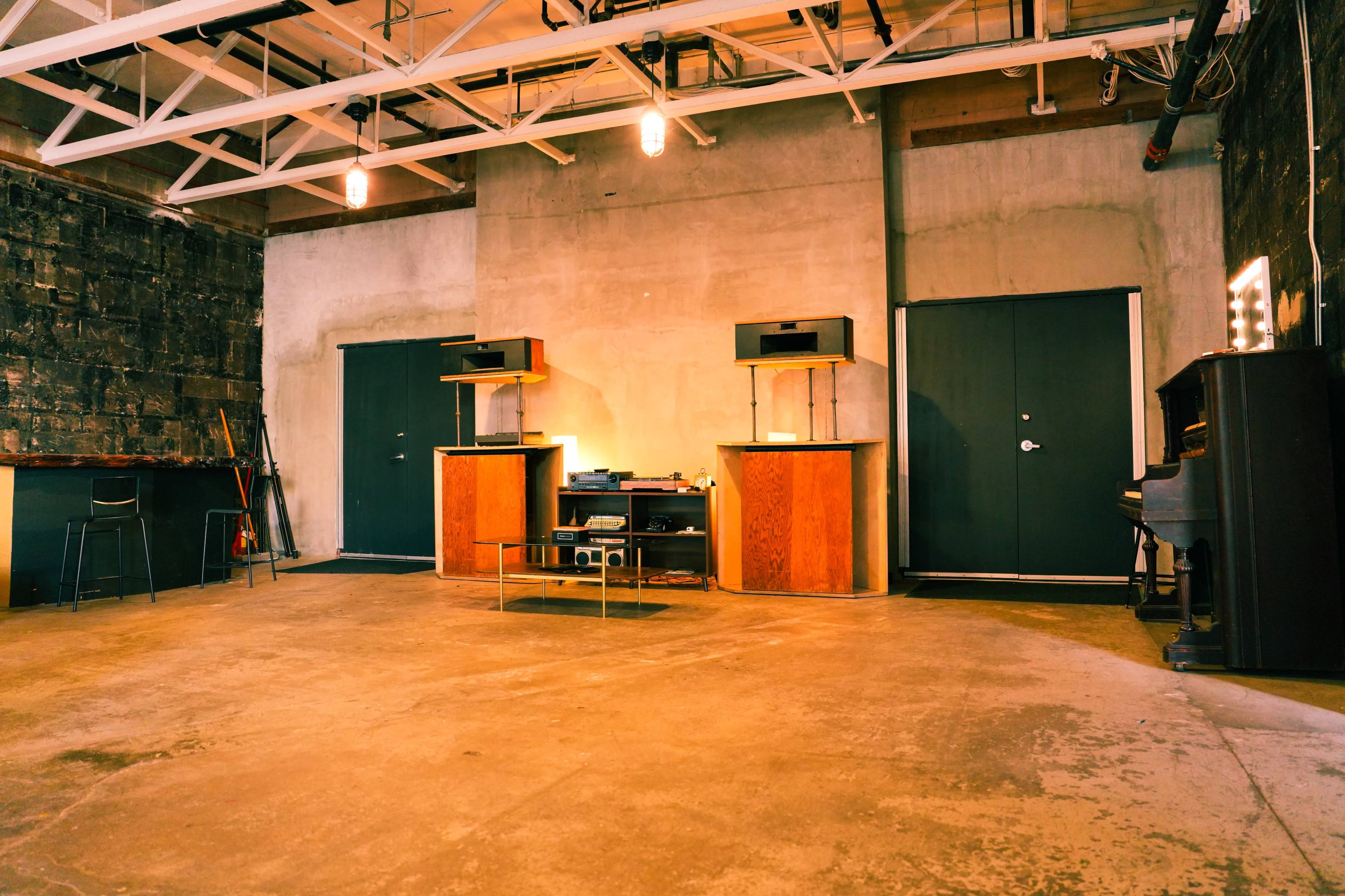The image shows an industrial-style room with a concrete floor, two dark green doors, a glass coffee table, and wooden pillars that support a ceiling with exposed beams.