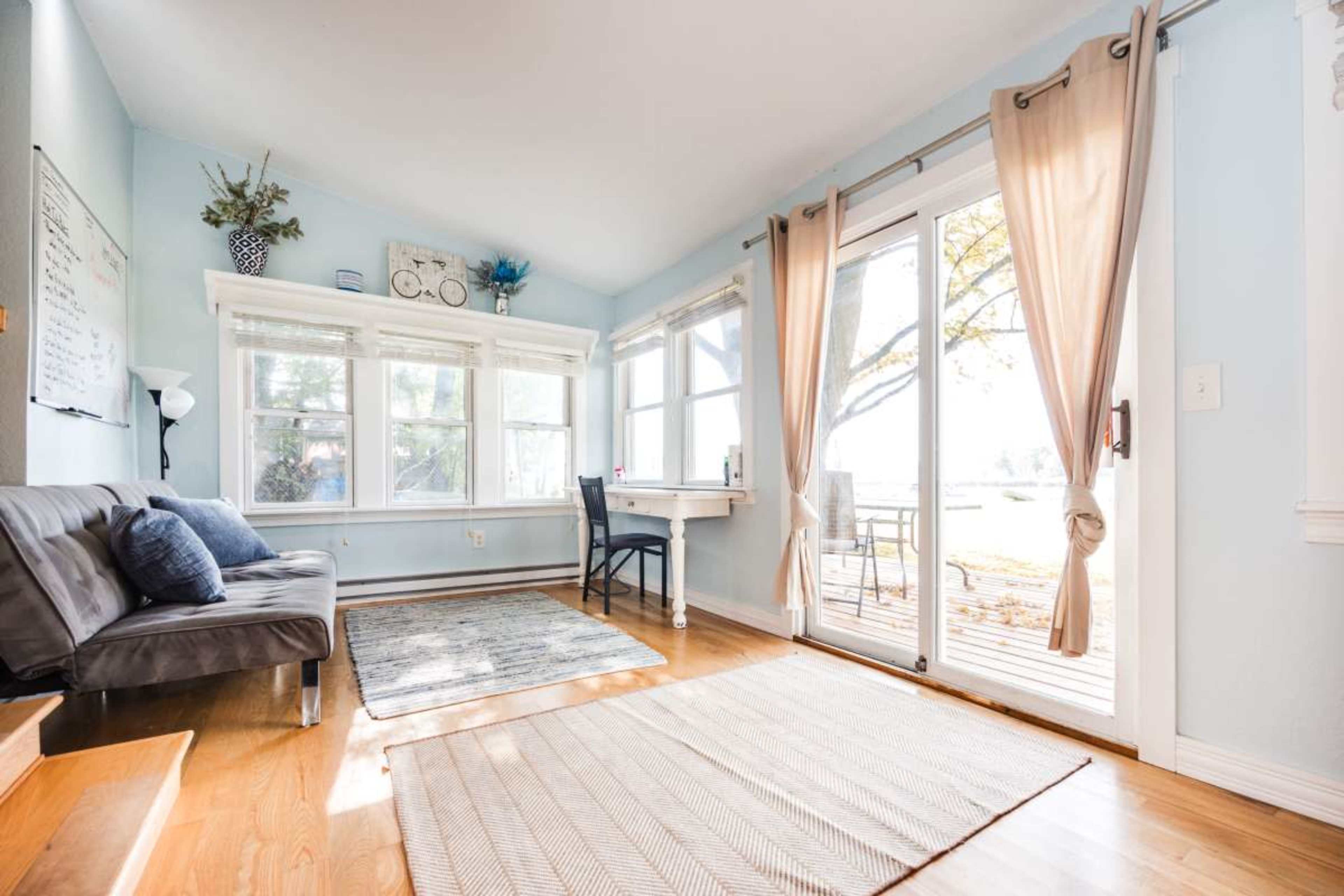 A cozy room features a gray sofa, a small desk by a window, and hardwood floors with a rug, allowing natural light to fill the space.