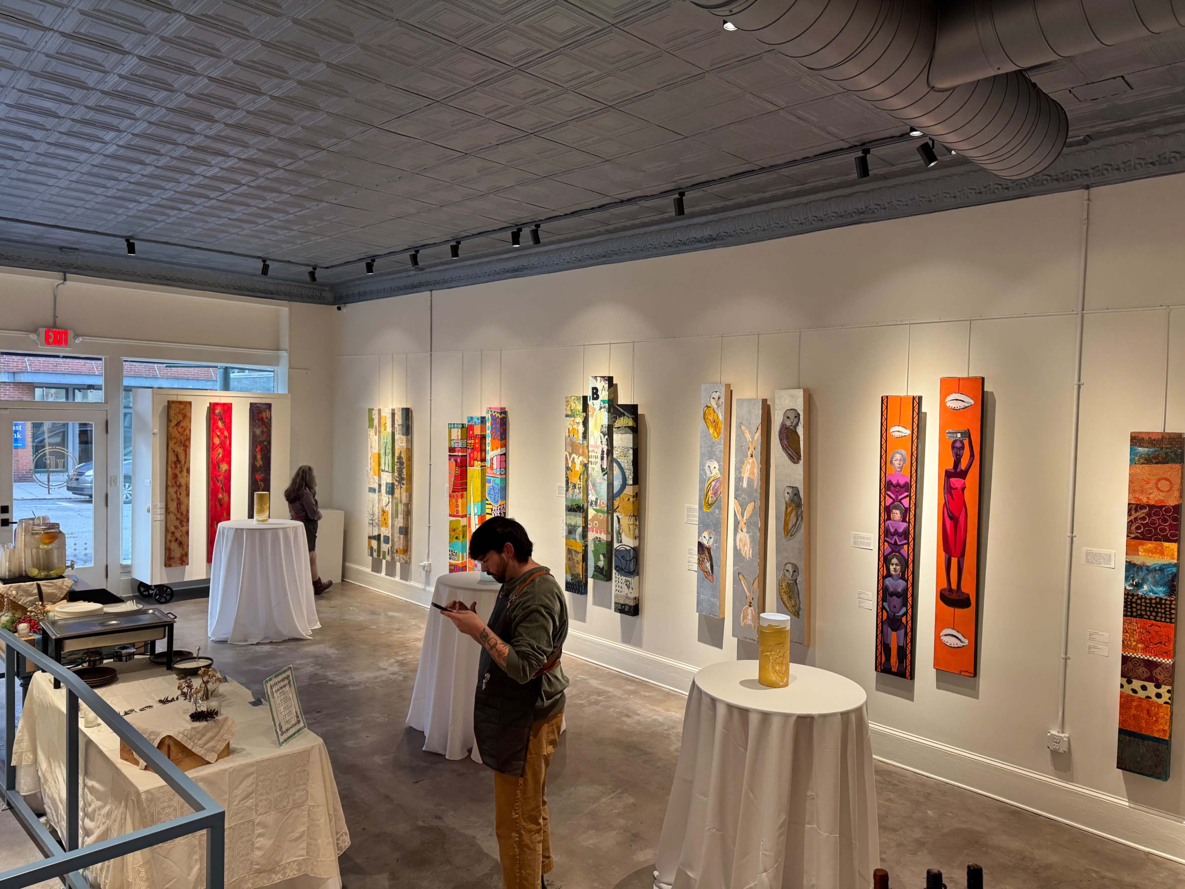 The image shows a gallery interior with colorful vertical paintings displayed on white walls, and two individuals engaged in activities at small tables.