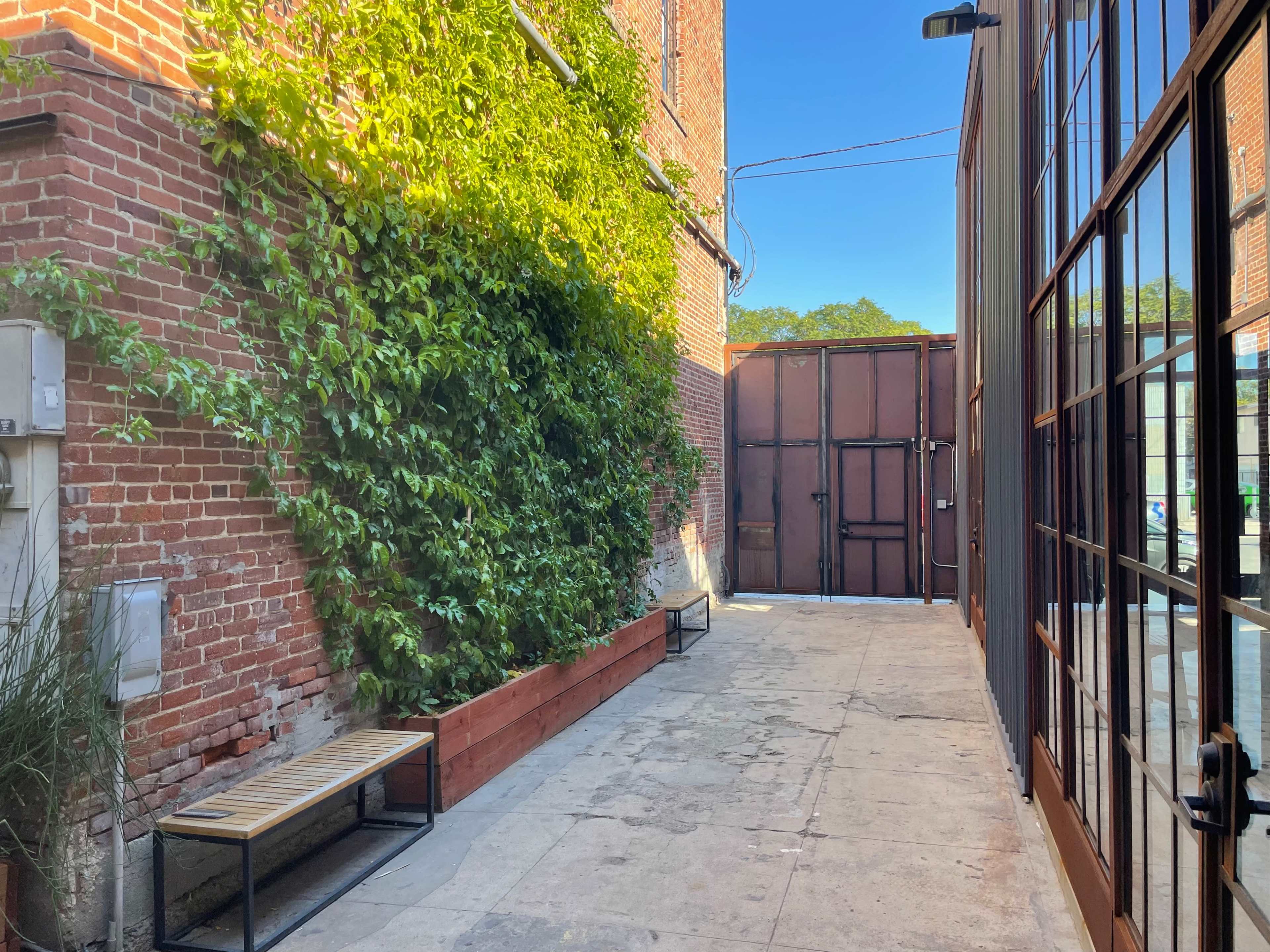 A narrow alleyway features a brick wall with greenery, benches on either side, and a large metal gate at the far end.