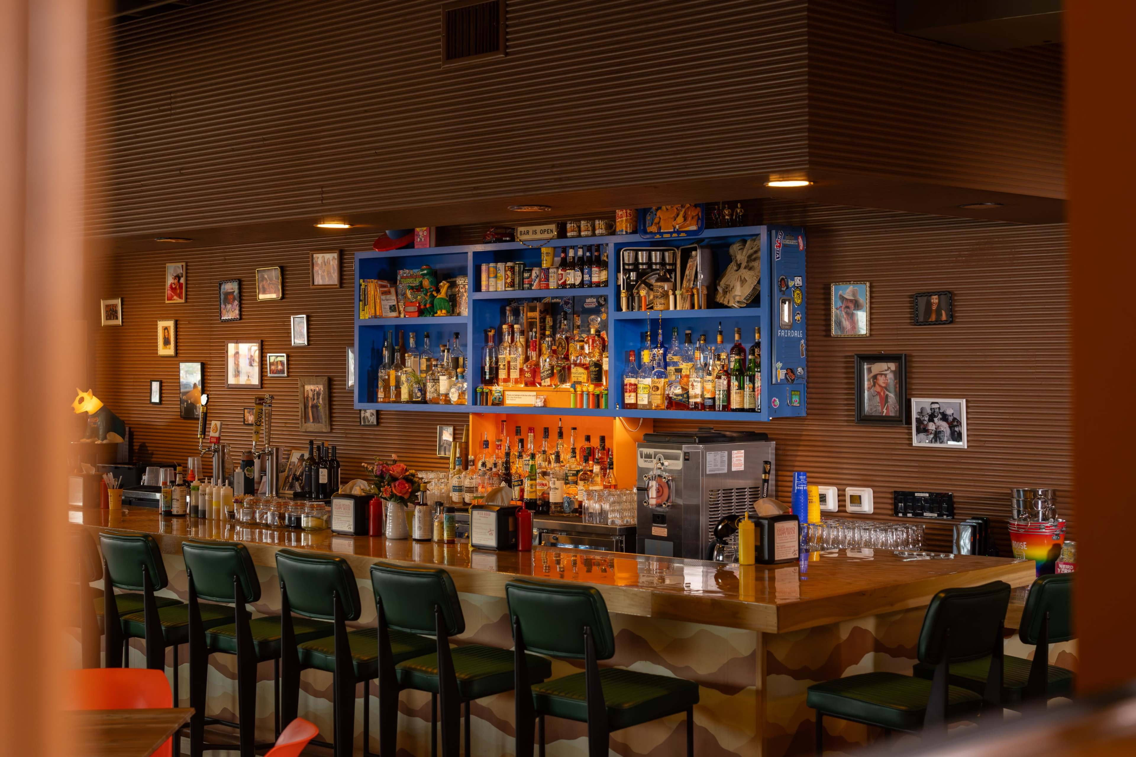 The image shows a bar with a wooden counter, several high chairs, and a colorful backlit shelf displaying various bottles of liquor and framed photos on the walls.