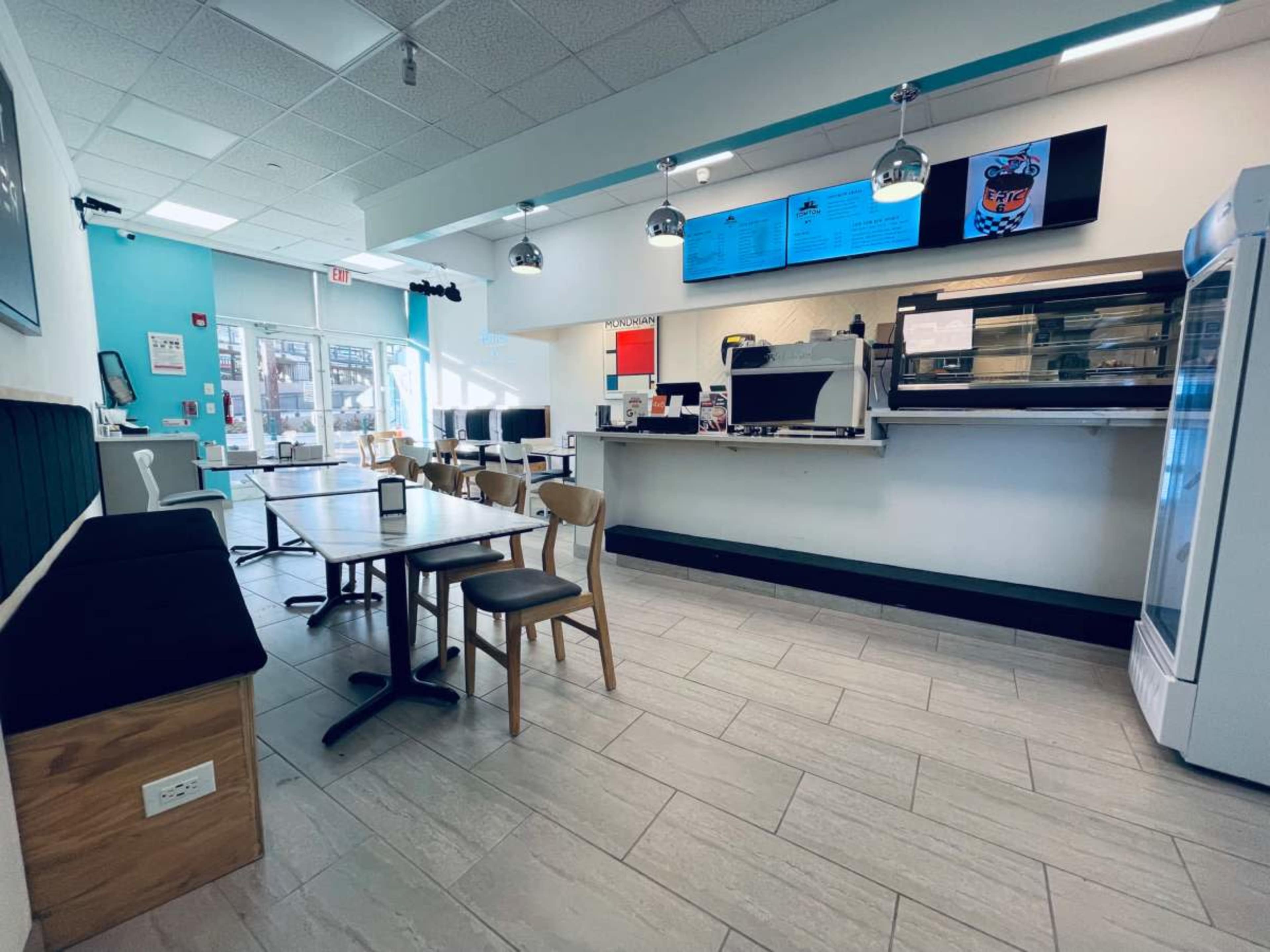 The image shows a modern eatery with a counter, seating area with tables and chairs, and digital screens displaying the menu.
