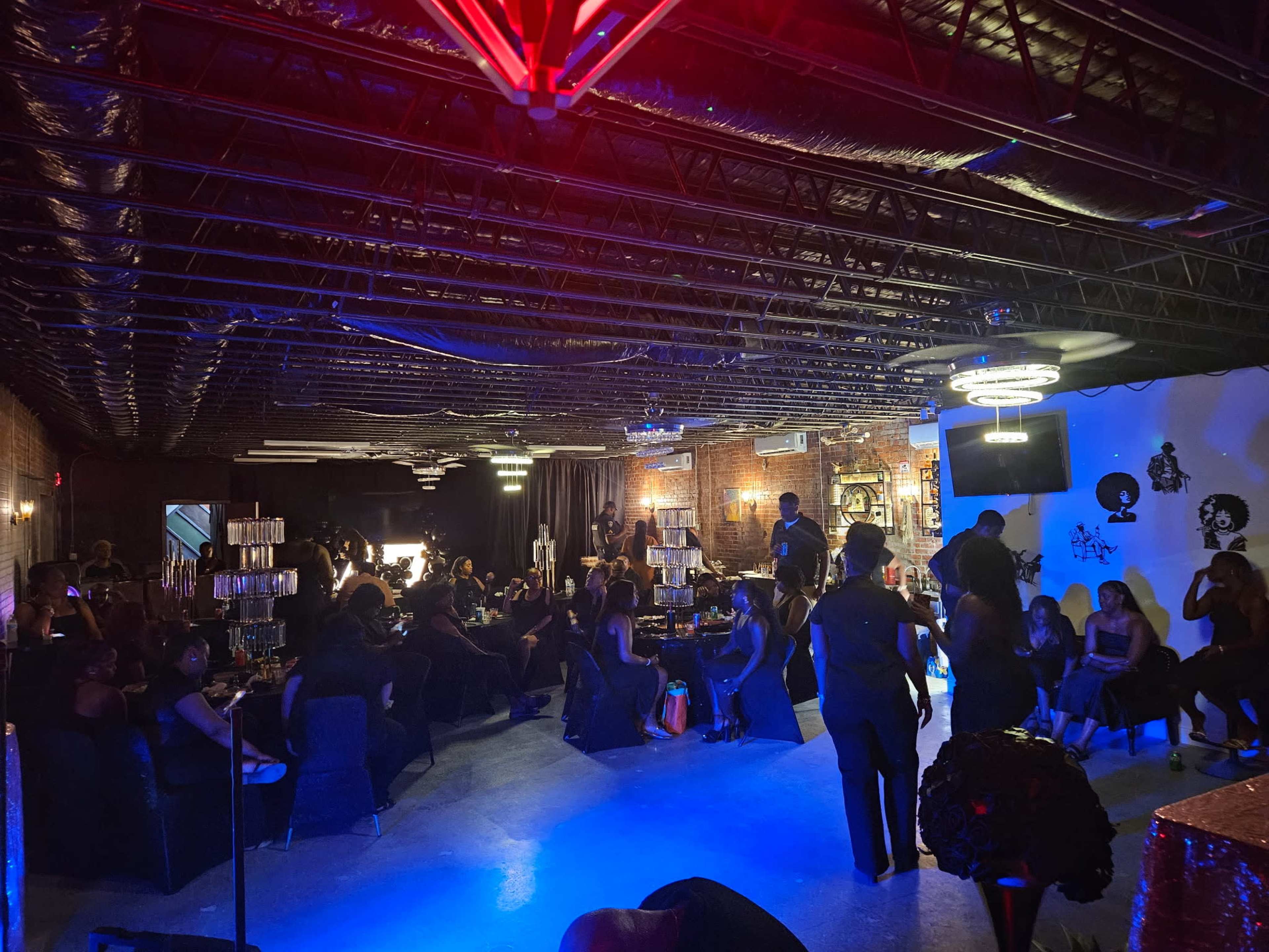 A crowded event space features numerous people mingling and seated in a dimly lit area with exposed brick walls and decorative lighting.