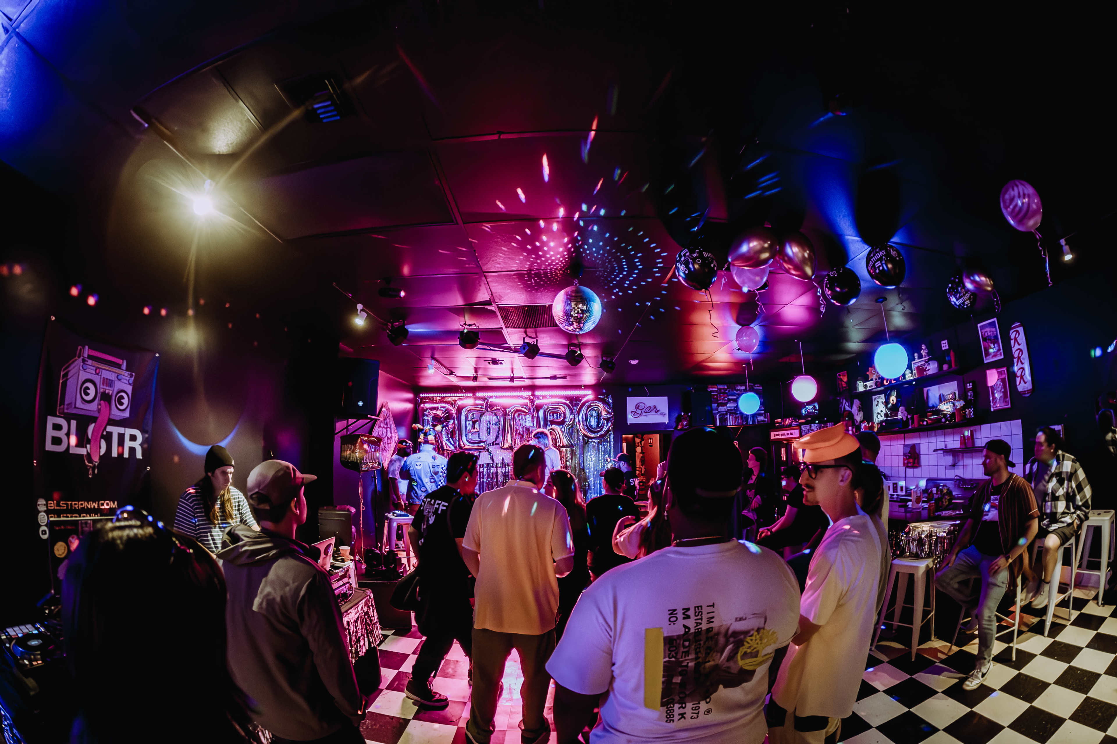 A lively gathering with people socializing and dancing in a colorful venue featuring disco lights and a checkered floor.