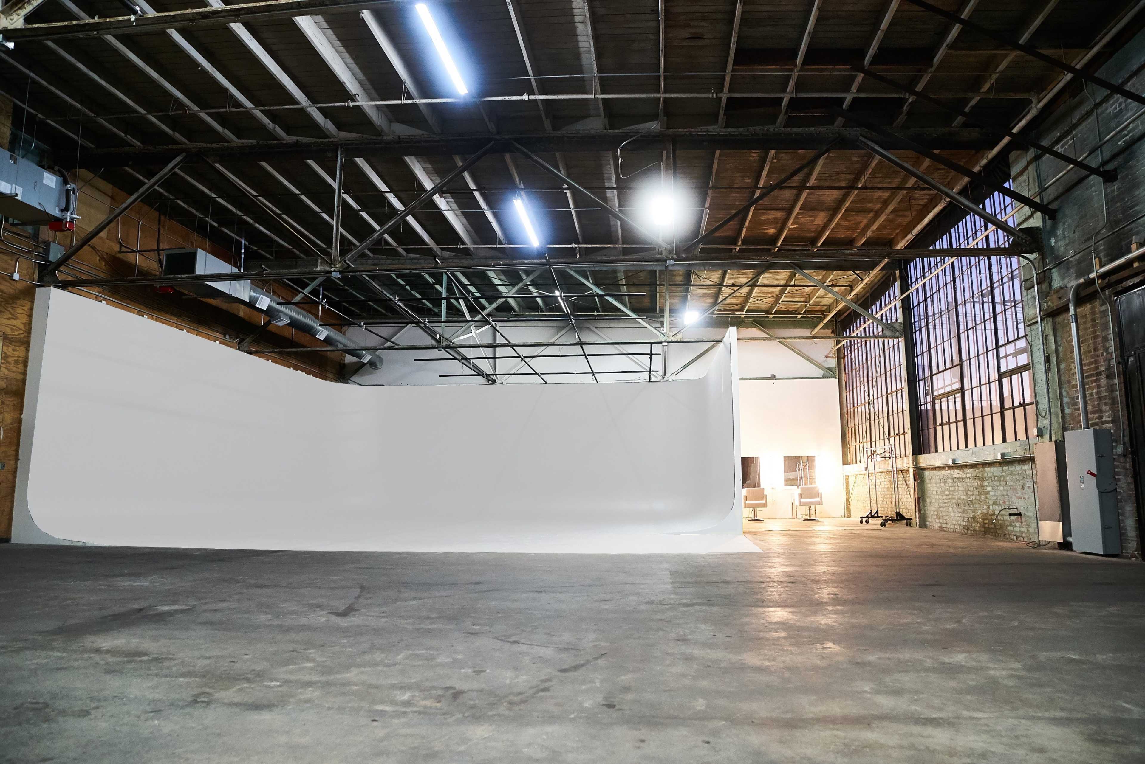 The image shows a spacious industrial studio with a white backdrop, large windows, and an empty concrete floor.