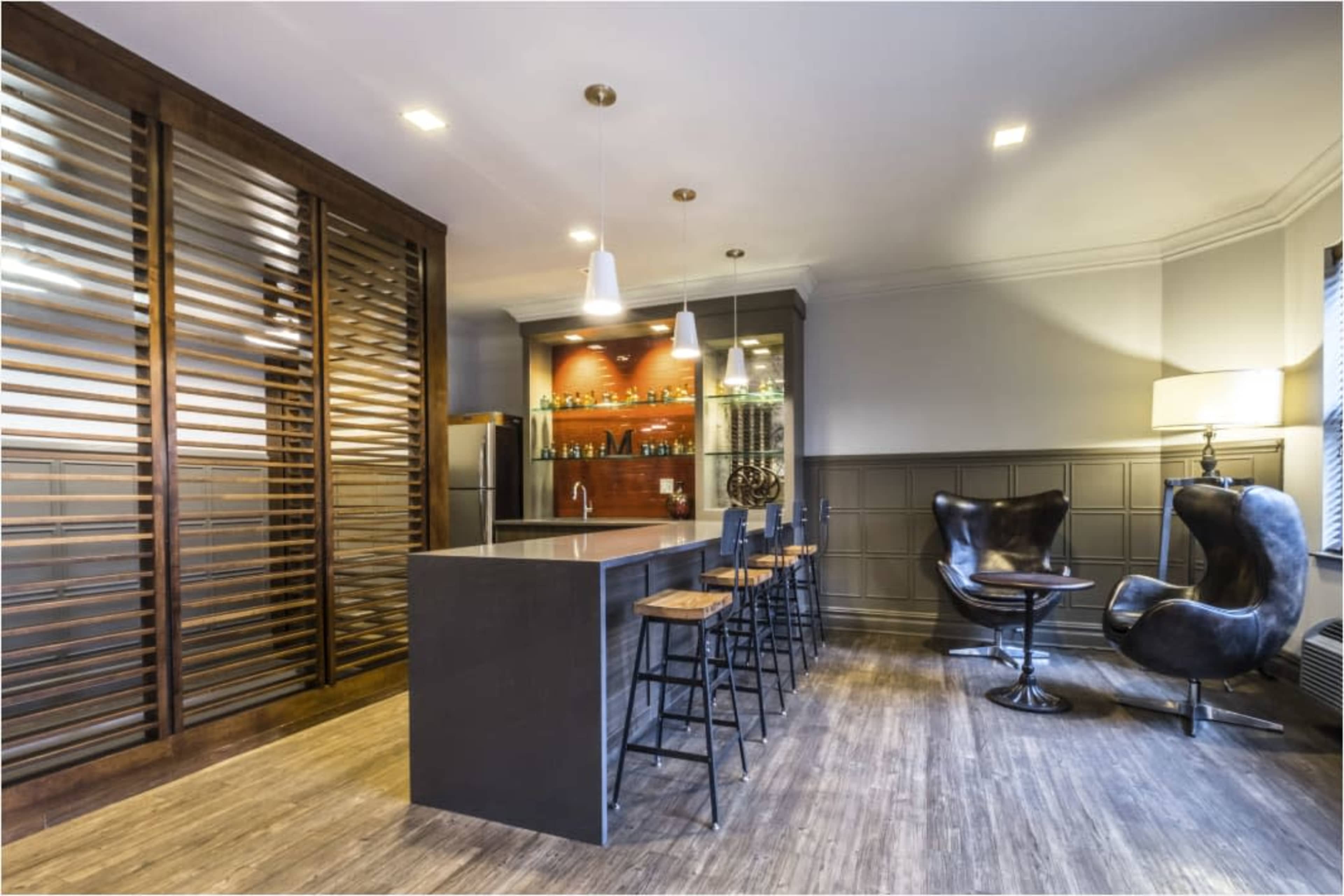 The image shows a modern interior space featuring a sleek bar with stools, a well-lit display of bottles, and two distinctive black chairs.