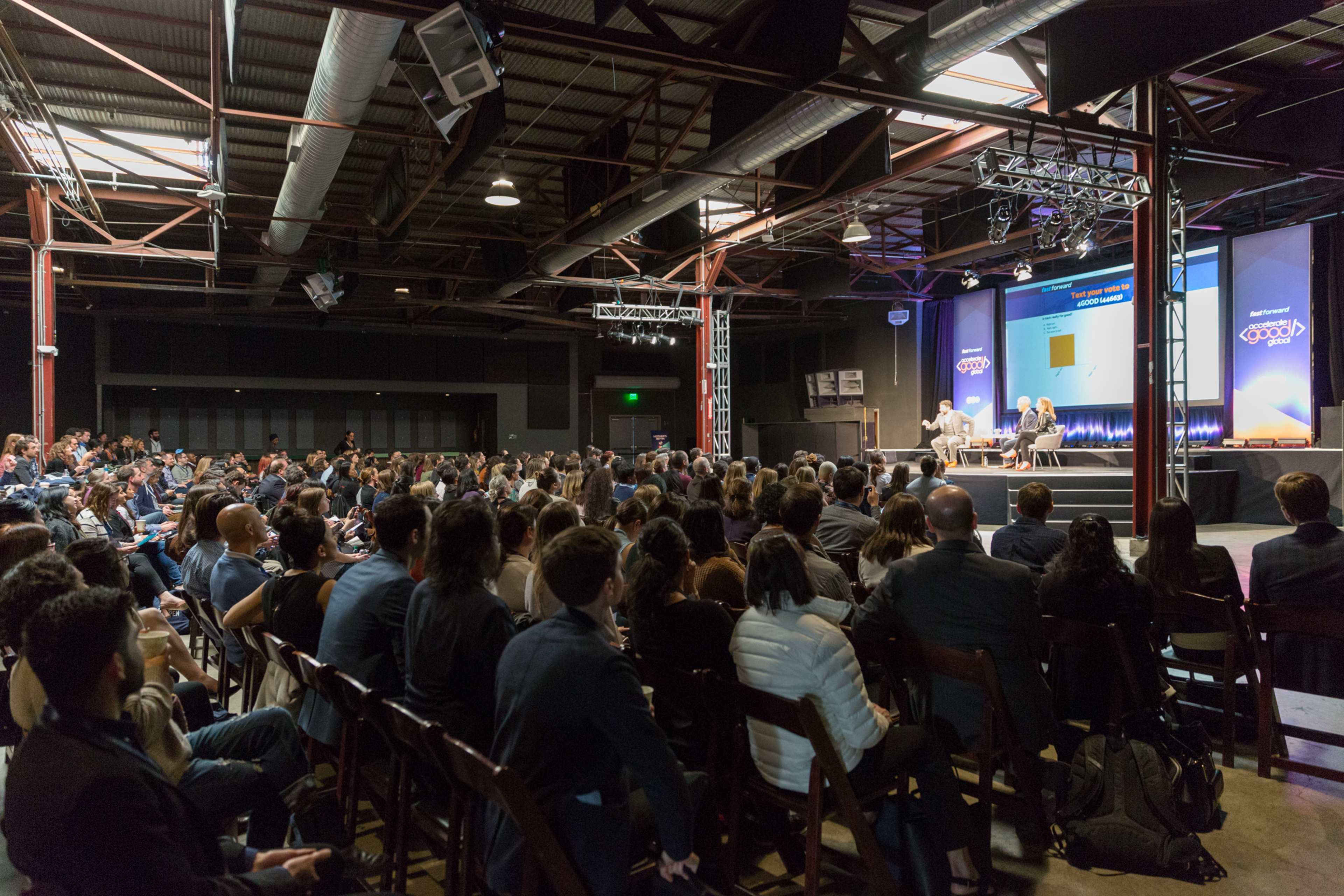 A large audience sits in a dimly lit industrial venue, facing a stage where two speakers are engaged in a discussion.