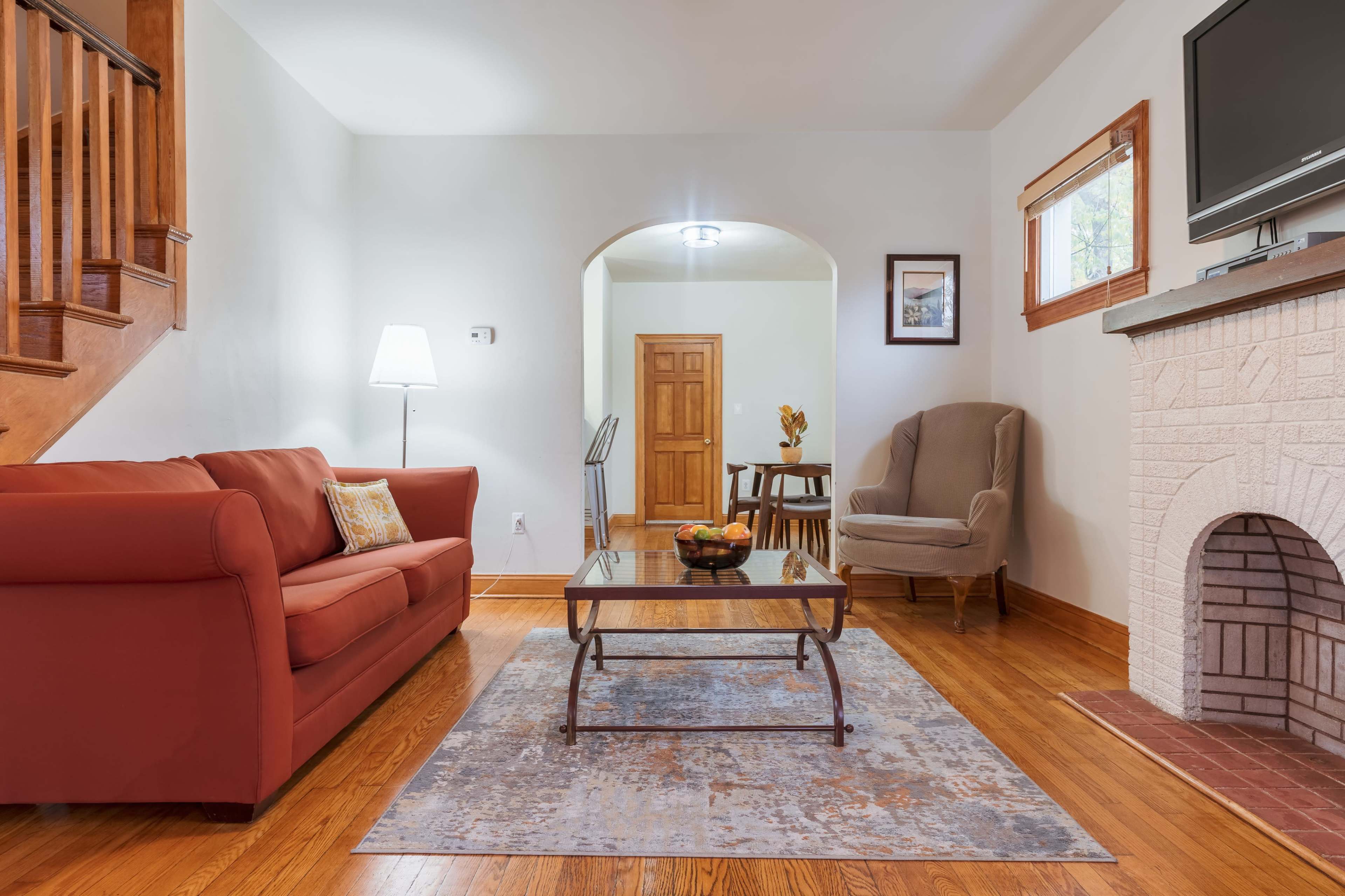 The image shows a living room with a red sofa, a coffee table, a beige armchair, and a fireplace, featuring hardwood floors and an archway leading to another room.
