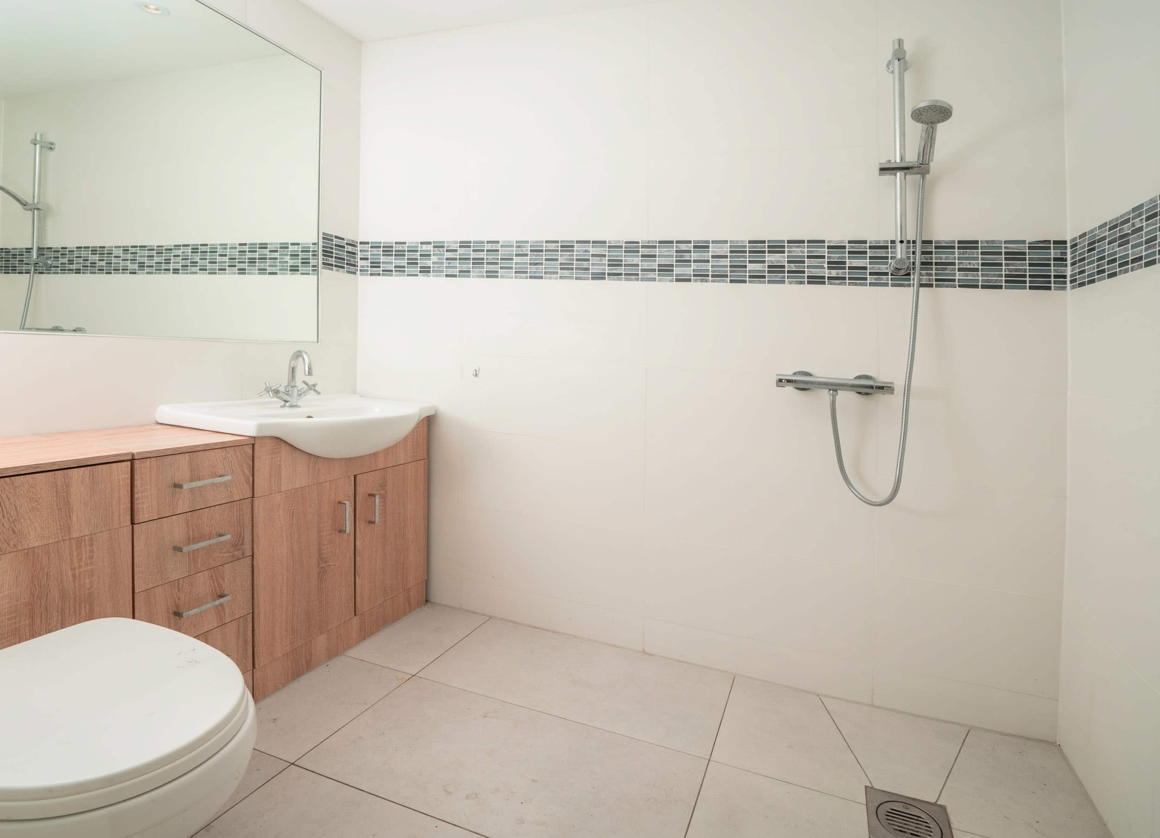 A clean bathroom featuring a shower, sink, wooden cabinet, and tiled floor with a decorative border.