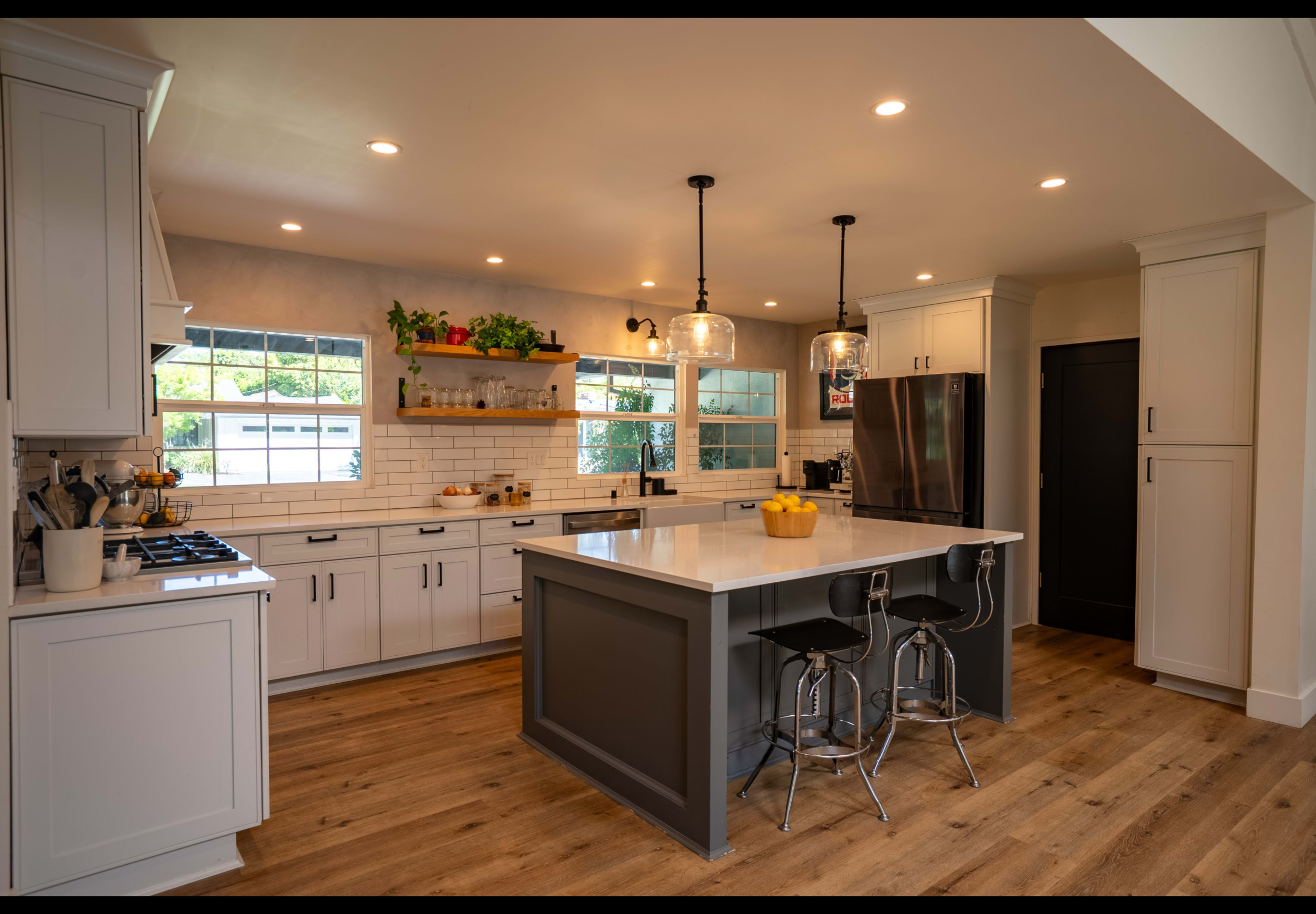 The kitchen features gray and white cabinetry, a central island with bar stools, and large windows allowing natural light.