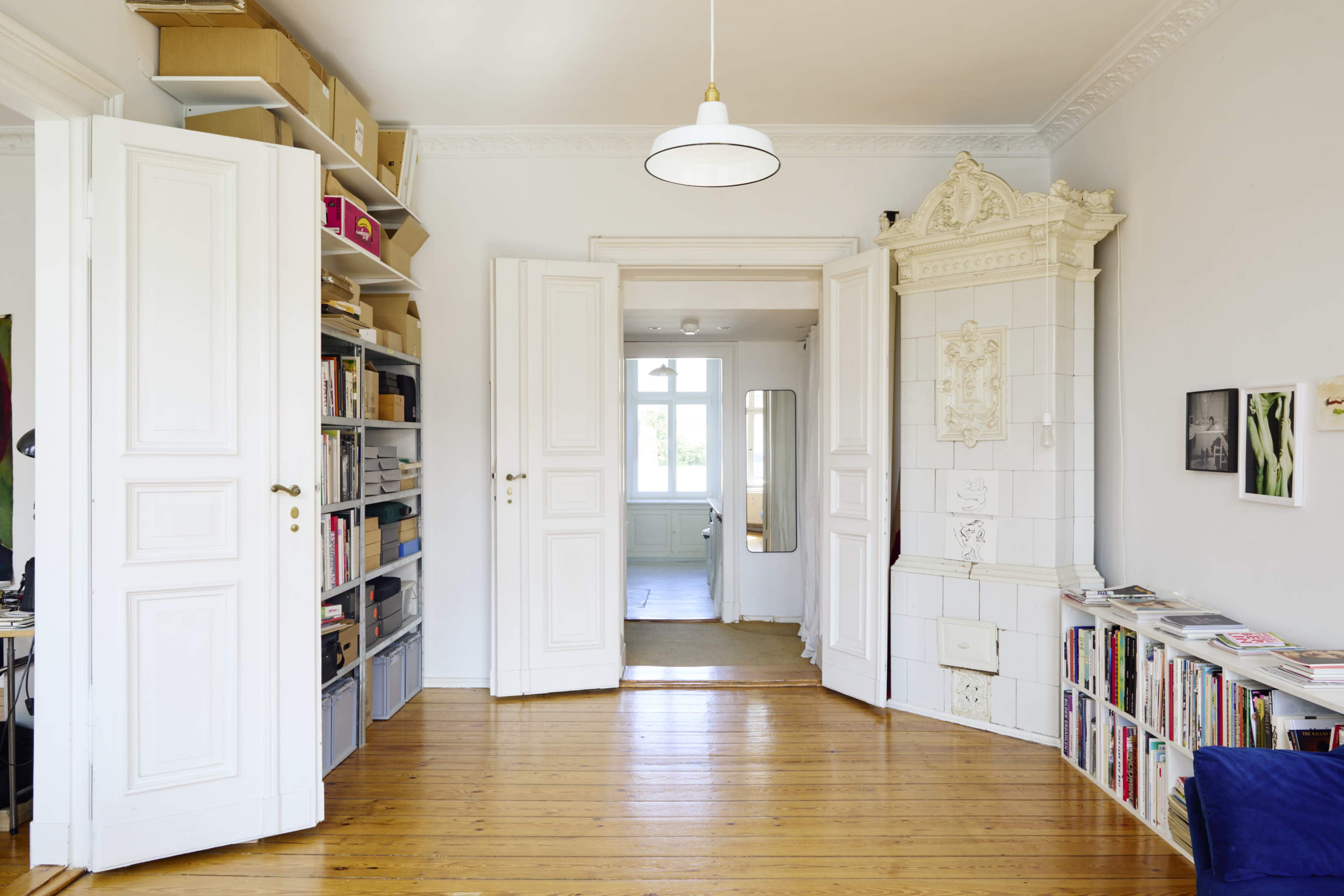 A brightly lit room features white double doors that open to another space, with wooden floors and shelves filled with books and storage boxes.