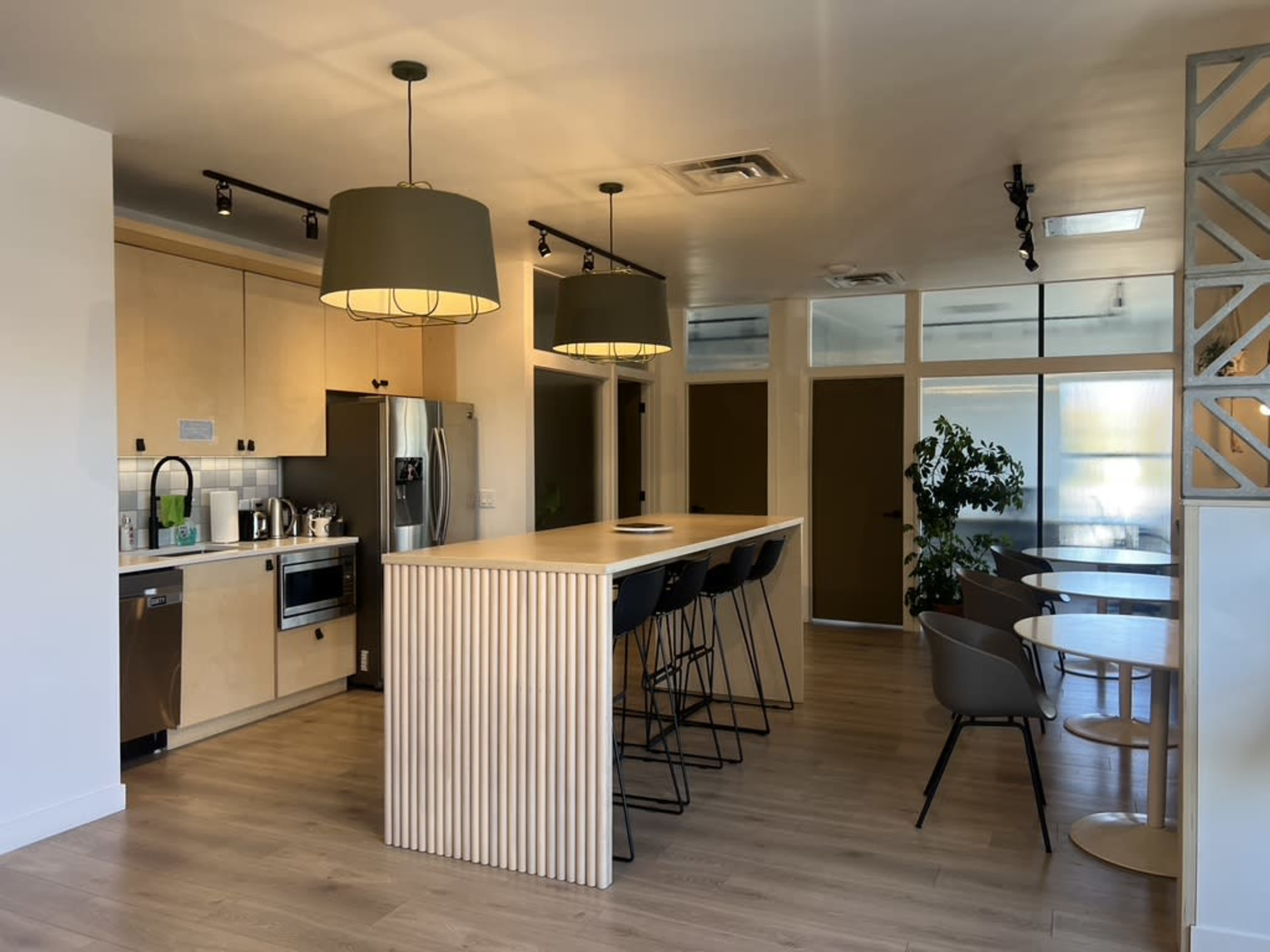 The image depicts a modern kitchen area with a large island and bar stools, featuring stainless steel appliances and ample natural light from large windows.