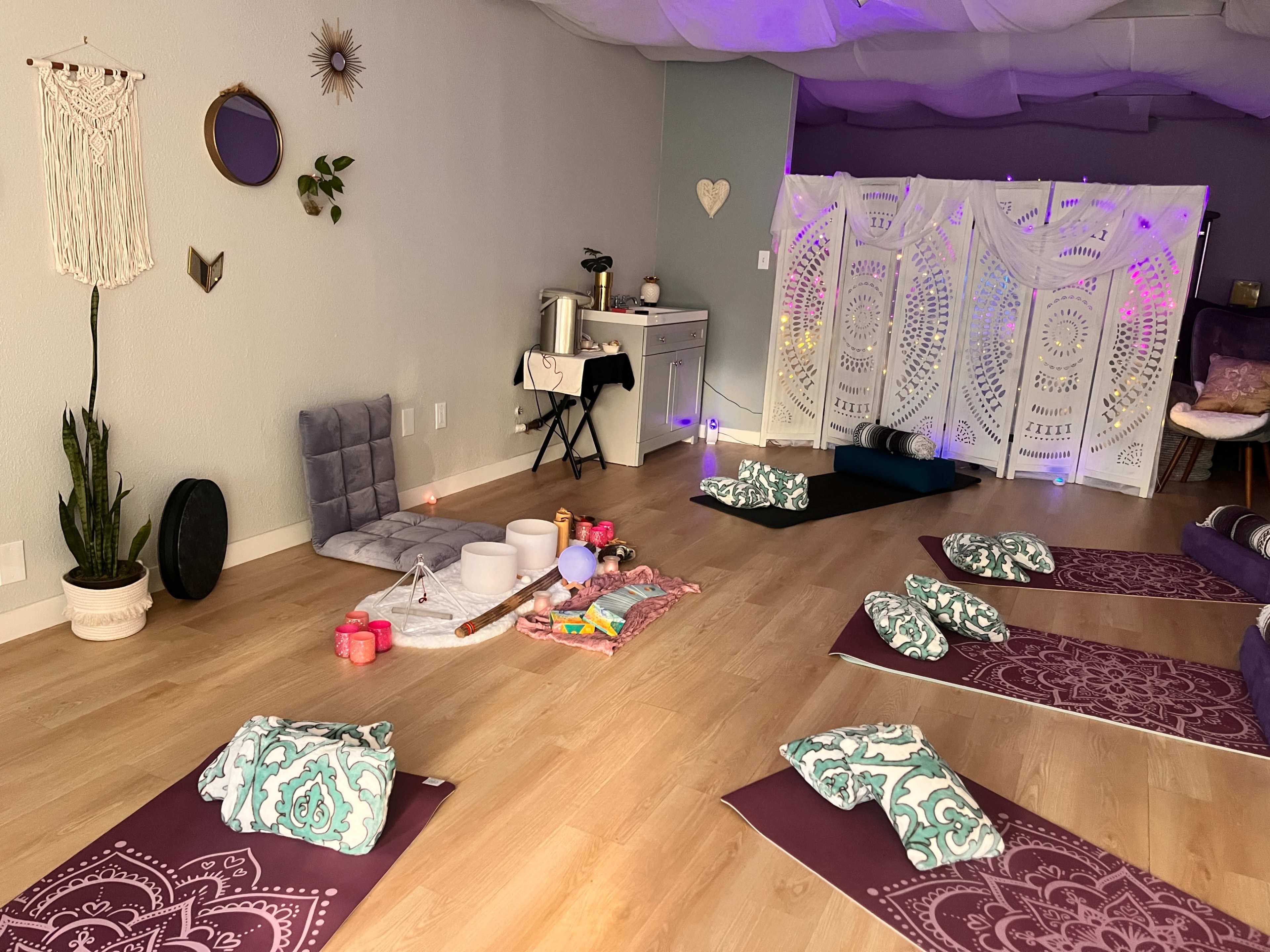 The image shows a serene yoga studio setup with mats aligned on the floor, various meditation tools arranged on a center cloth, and a backdrop of decorative screens illuminated with soft lighting.