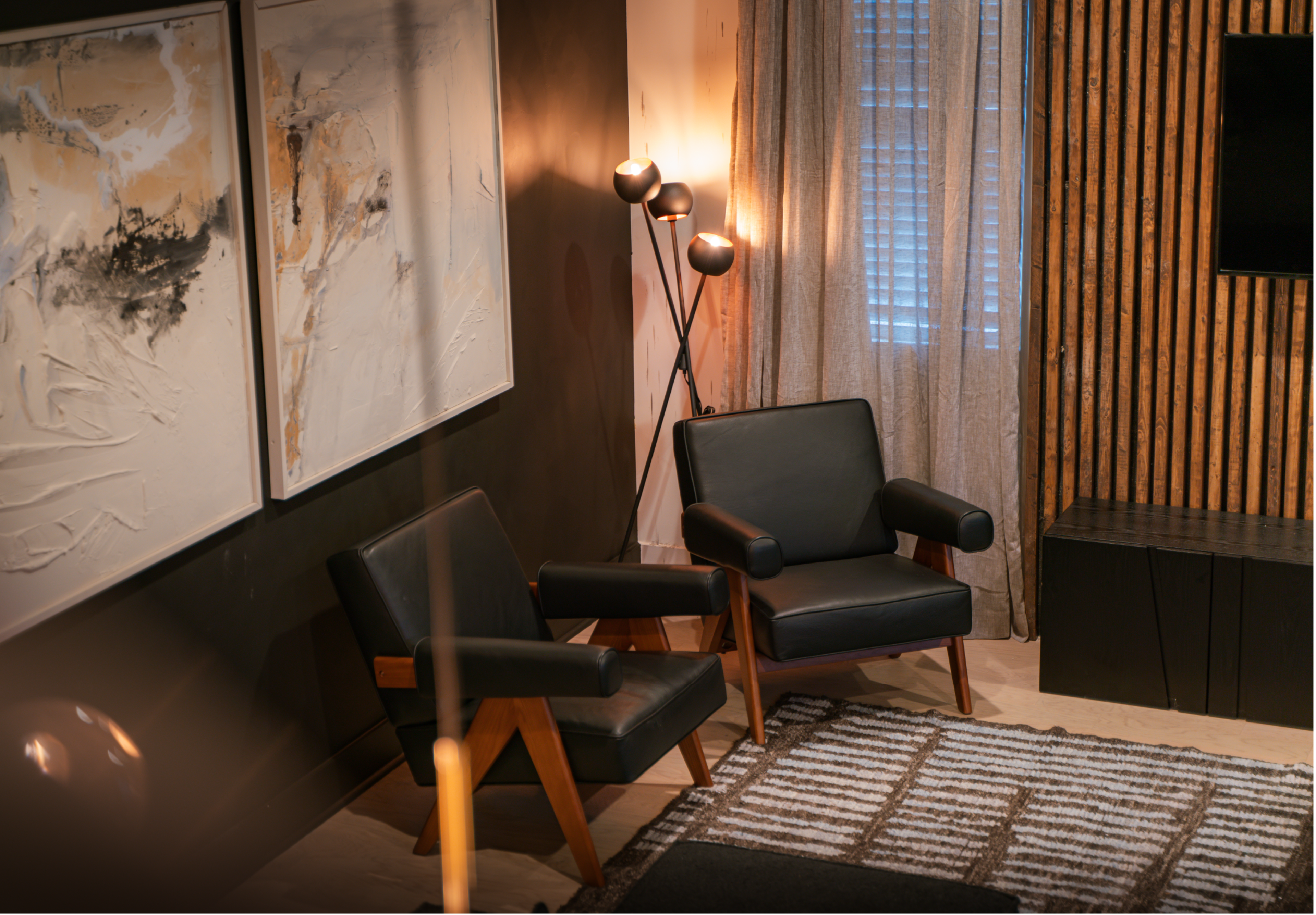 A modern living room with two black leather armchairs, a striped area rug, and abstract wall art, illuminated by a floor lamp and natural light filtering through sheer curtains.