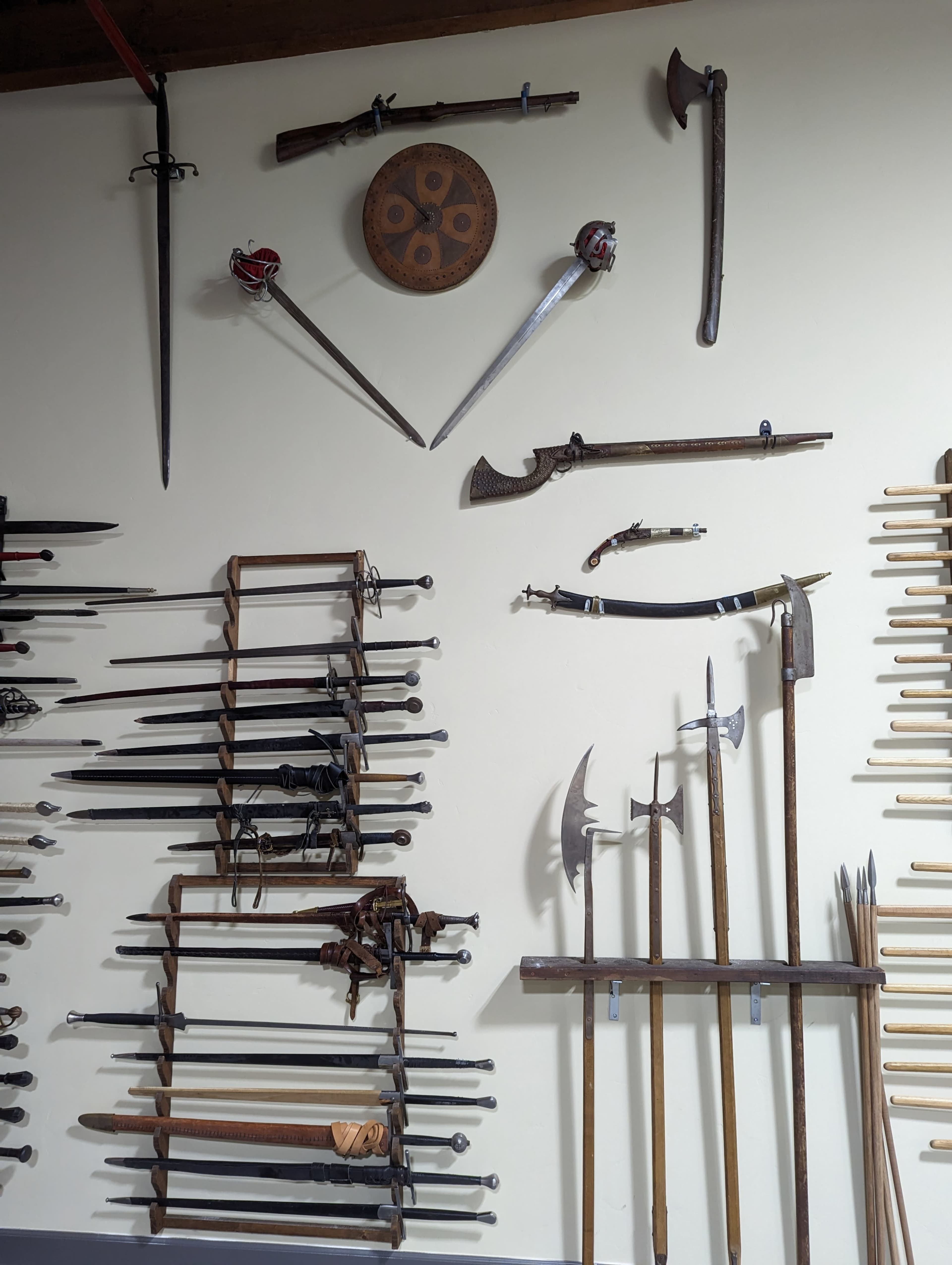 The image shows a wall display of various swords, axes, and bladed weapons arranged both vertically and horizontally, along with a round shield at the top center.