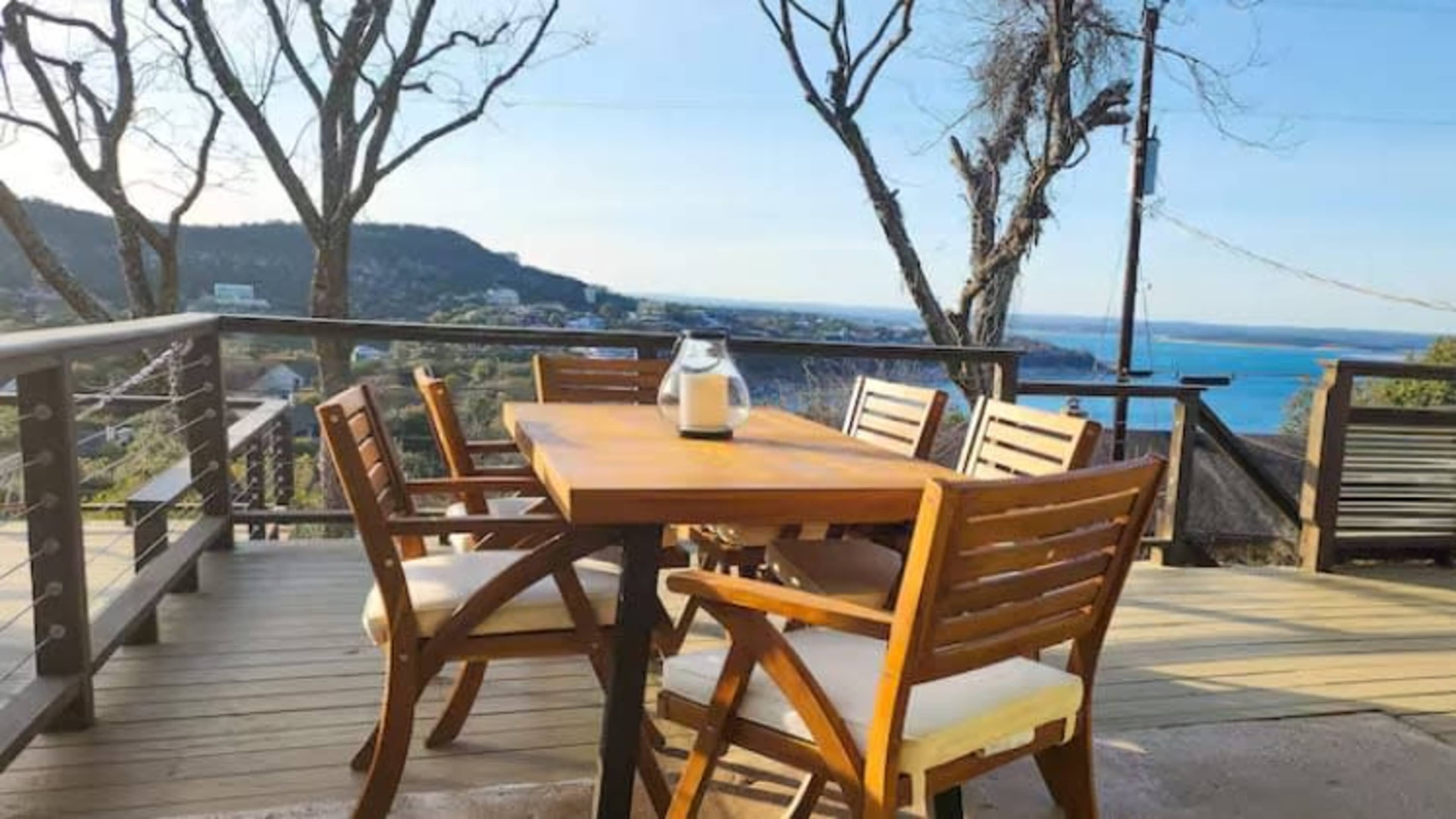 A wooden dining table with chairs is set on a deck overlooking a coastal view.