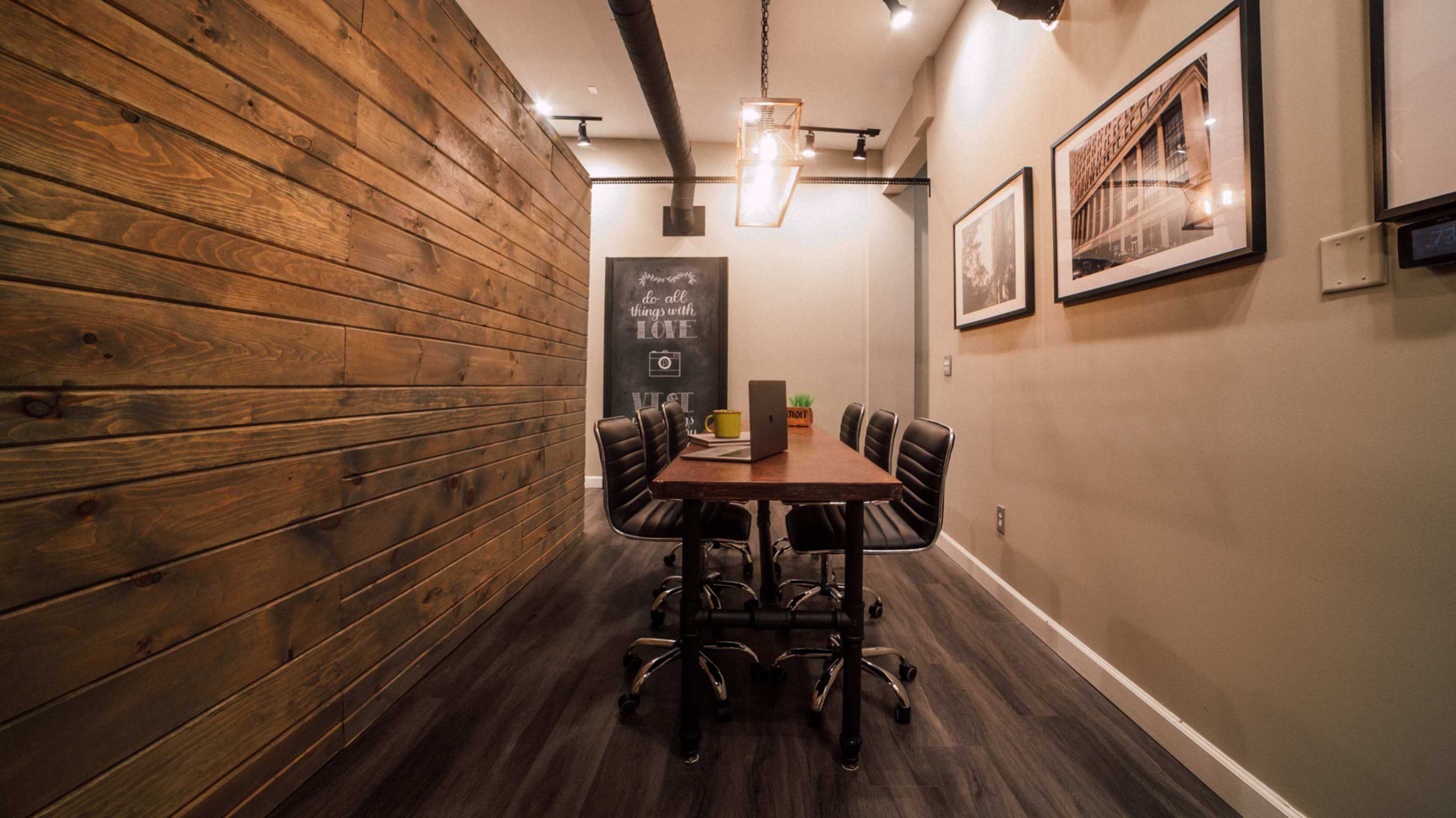 A narrow meeting room features a dark wood table surrounded by six black swivel chairs, with a textured wood wall and framed photographs adorning the walls.