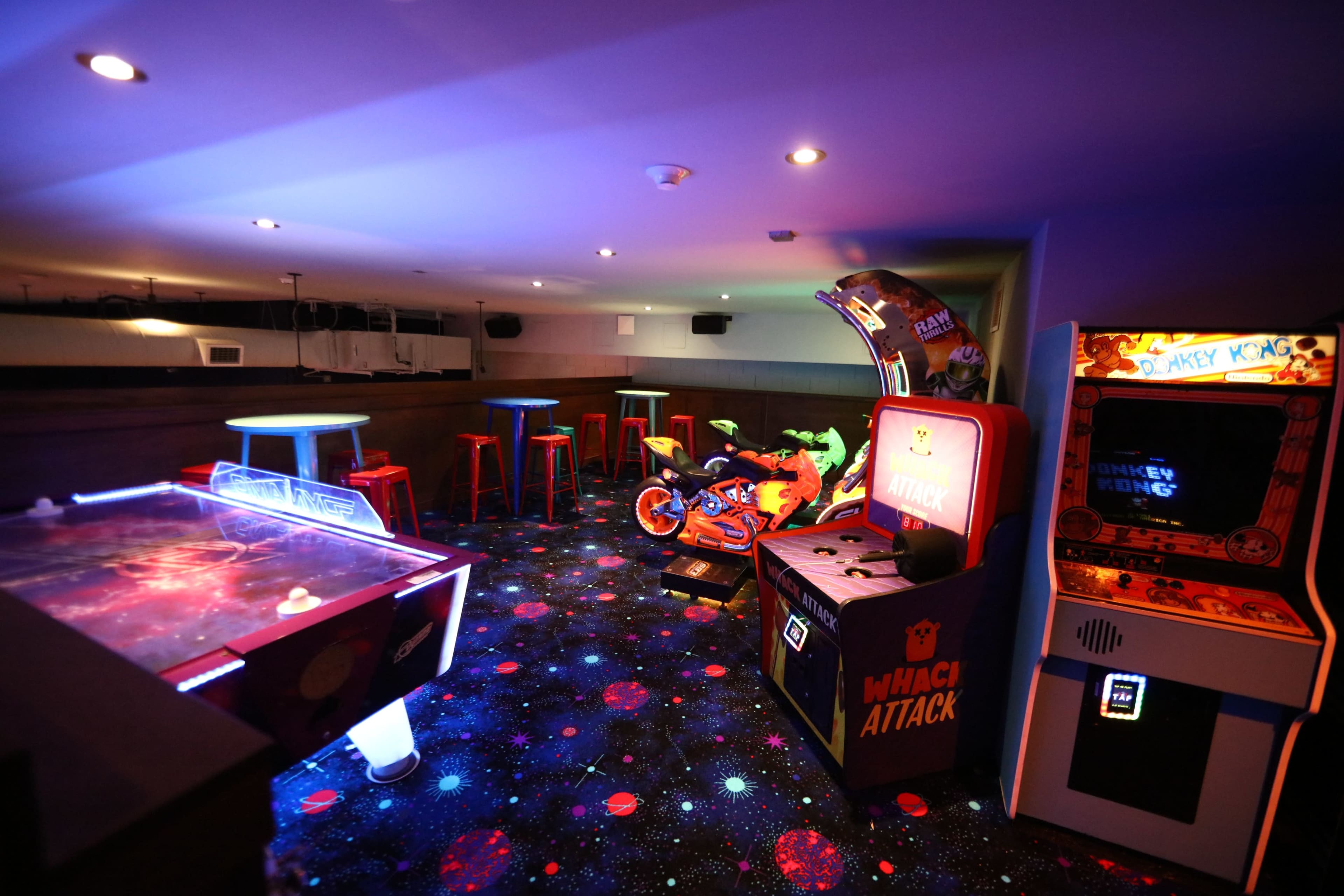 The image shows a dimly lit arcade room featuring various gaming machines, including an air hockey table and a motorcycle racing game, along with colorful seating and a starry carpet.