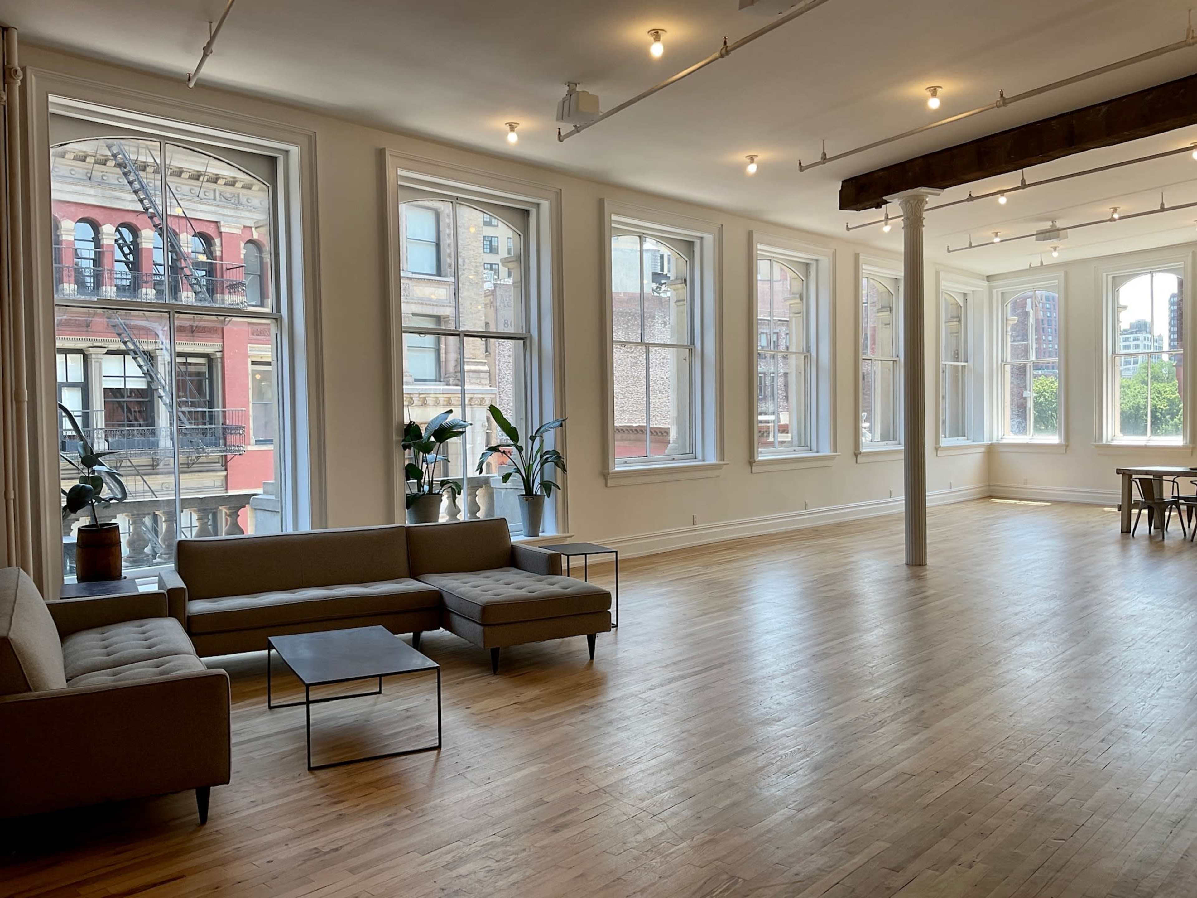 A spacious, well-lit room with large windows, wooden floors, and modern furniture, including sofas and a dining table.