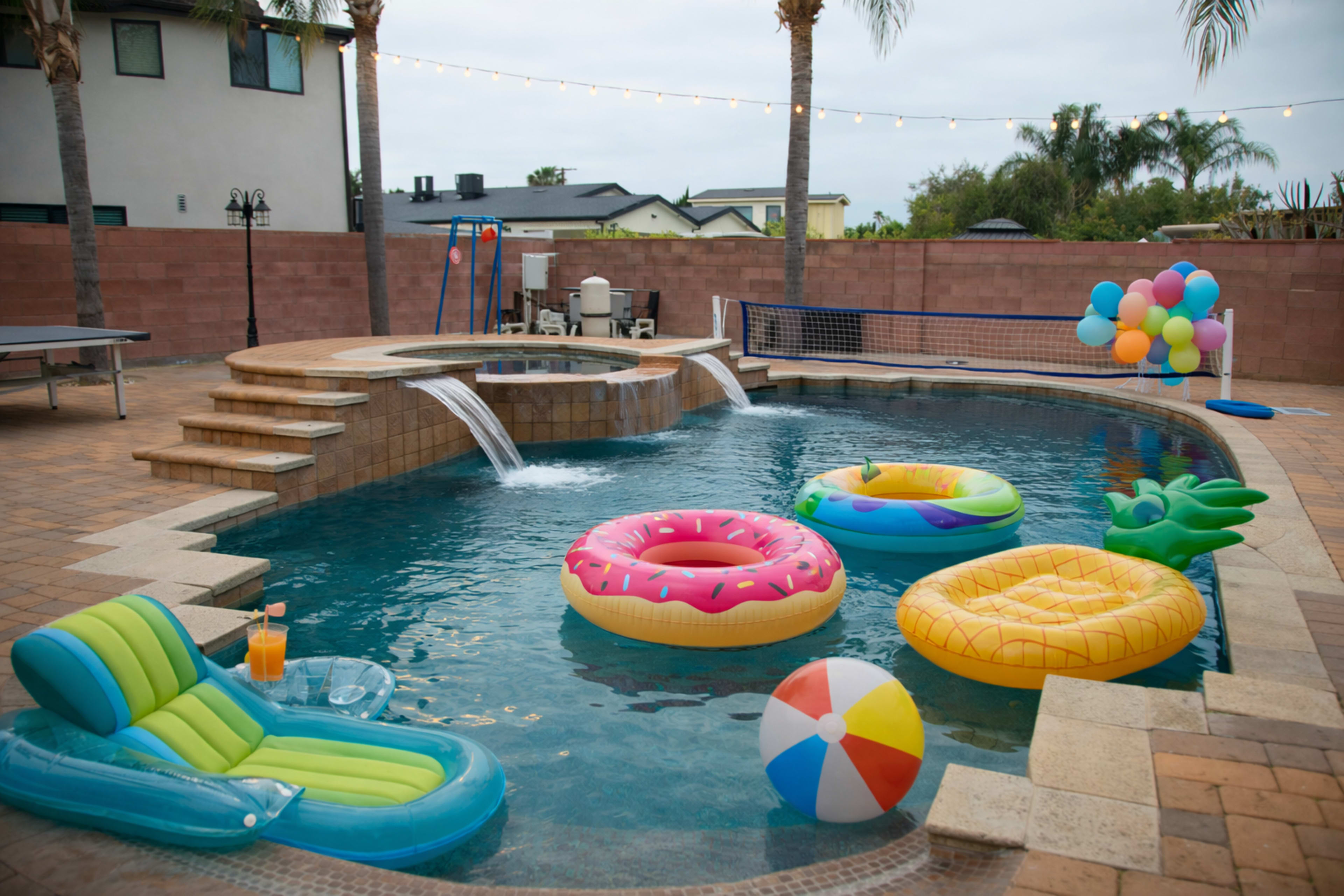 The scene shows a swimming pool area with colorful inflatable floats, a water feature, and a beach ball, surrounded by palm trees and a patio.