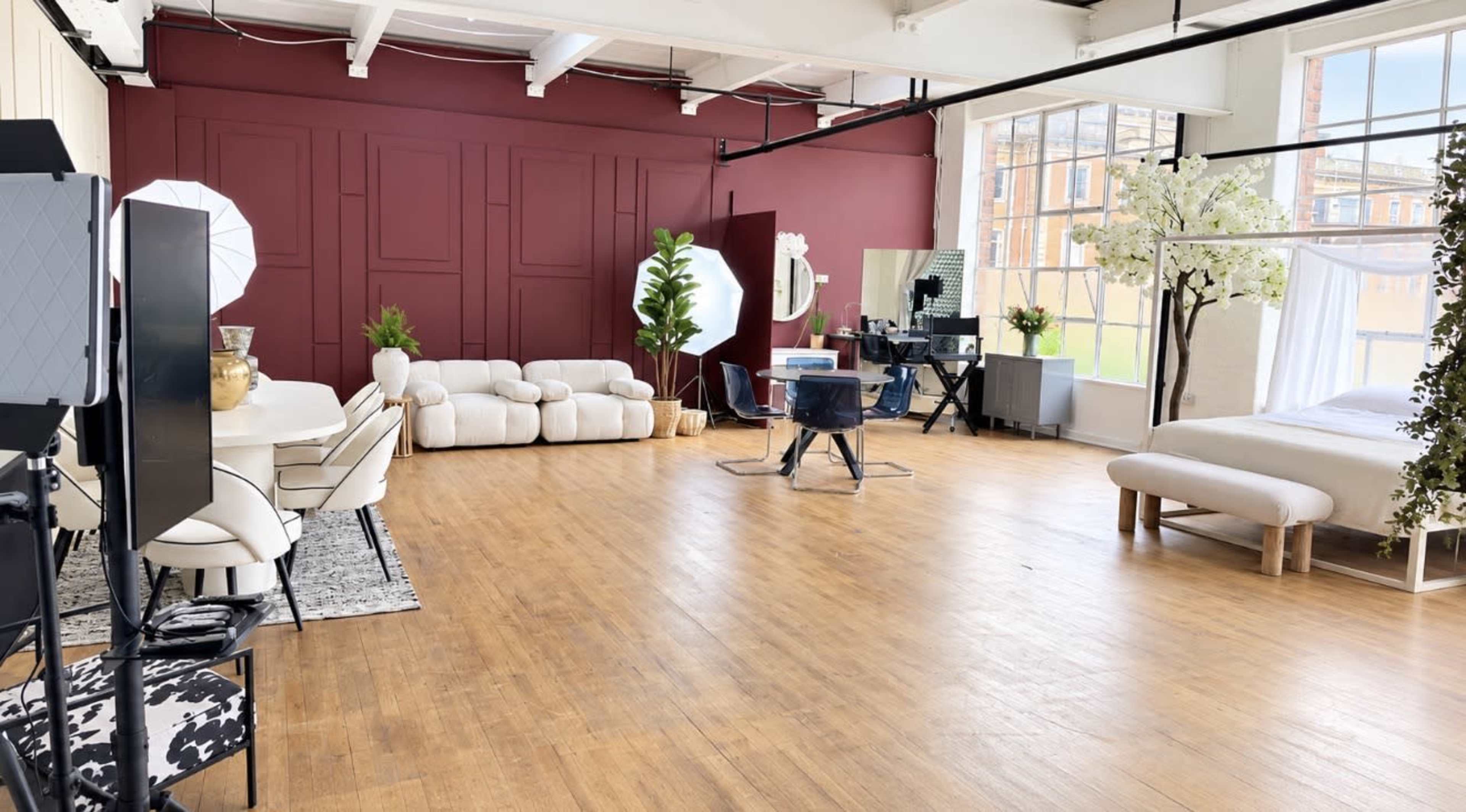 The image shows a bright, spacious loft with wooden flooring, featuring a white dining table set, a lounge area with plush seating, and a workspace with mirrors and styling chairs.