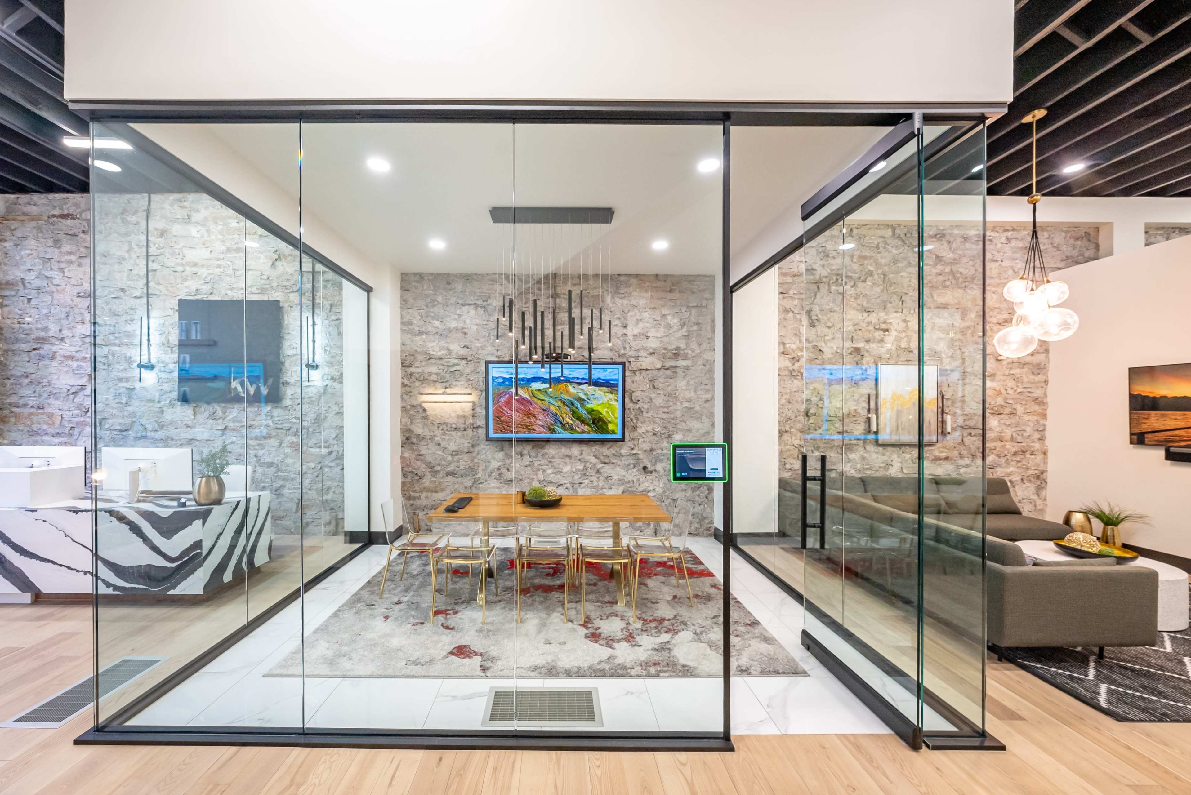 A modern dining area enclosed in glass, featuring a wooden table surrounded by transparent chairs, with stone walls and artwork in the background.