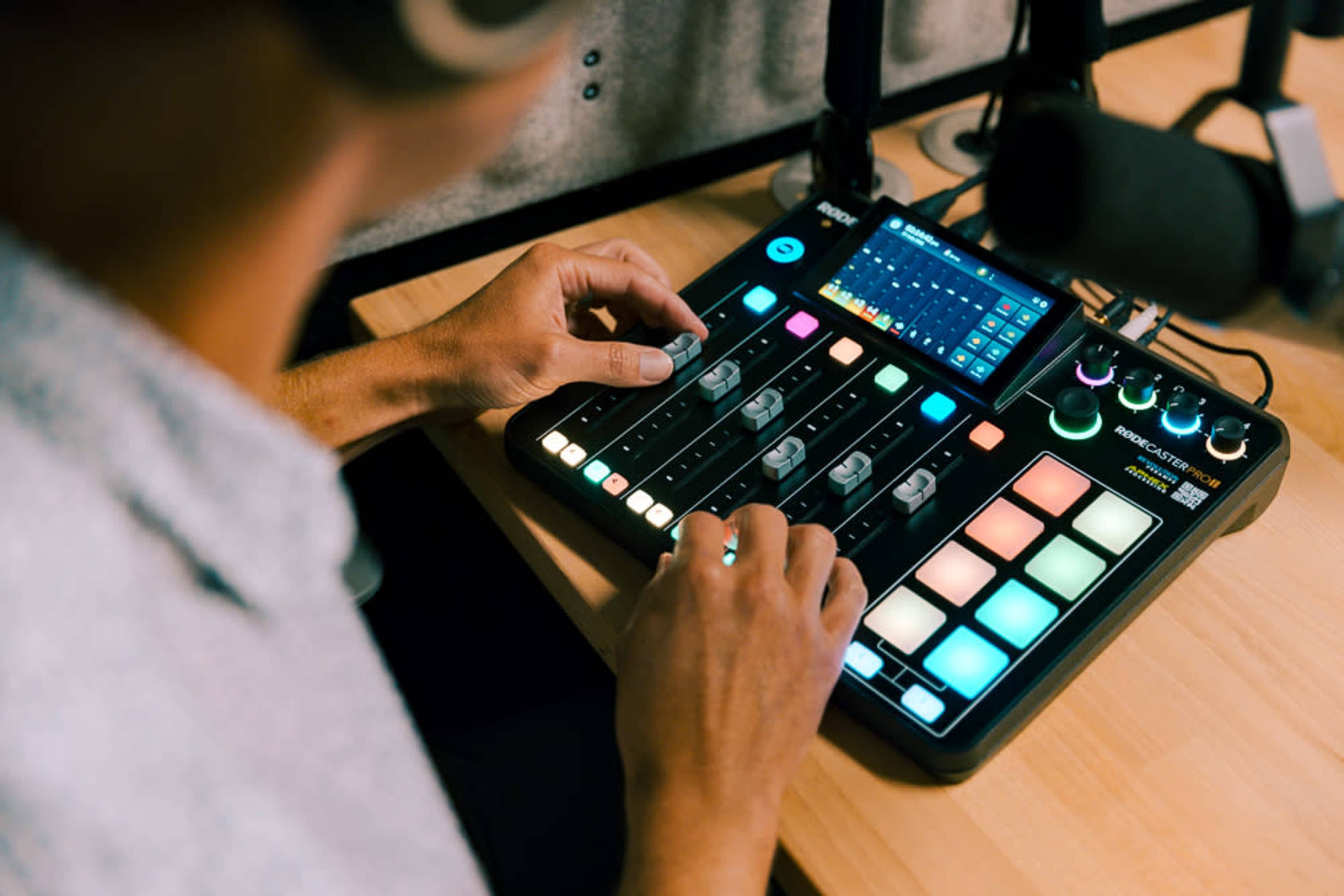 A person operates a digital audio mixer with multiple sliders and colorful buttons on a wooden surface.