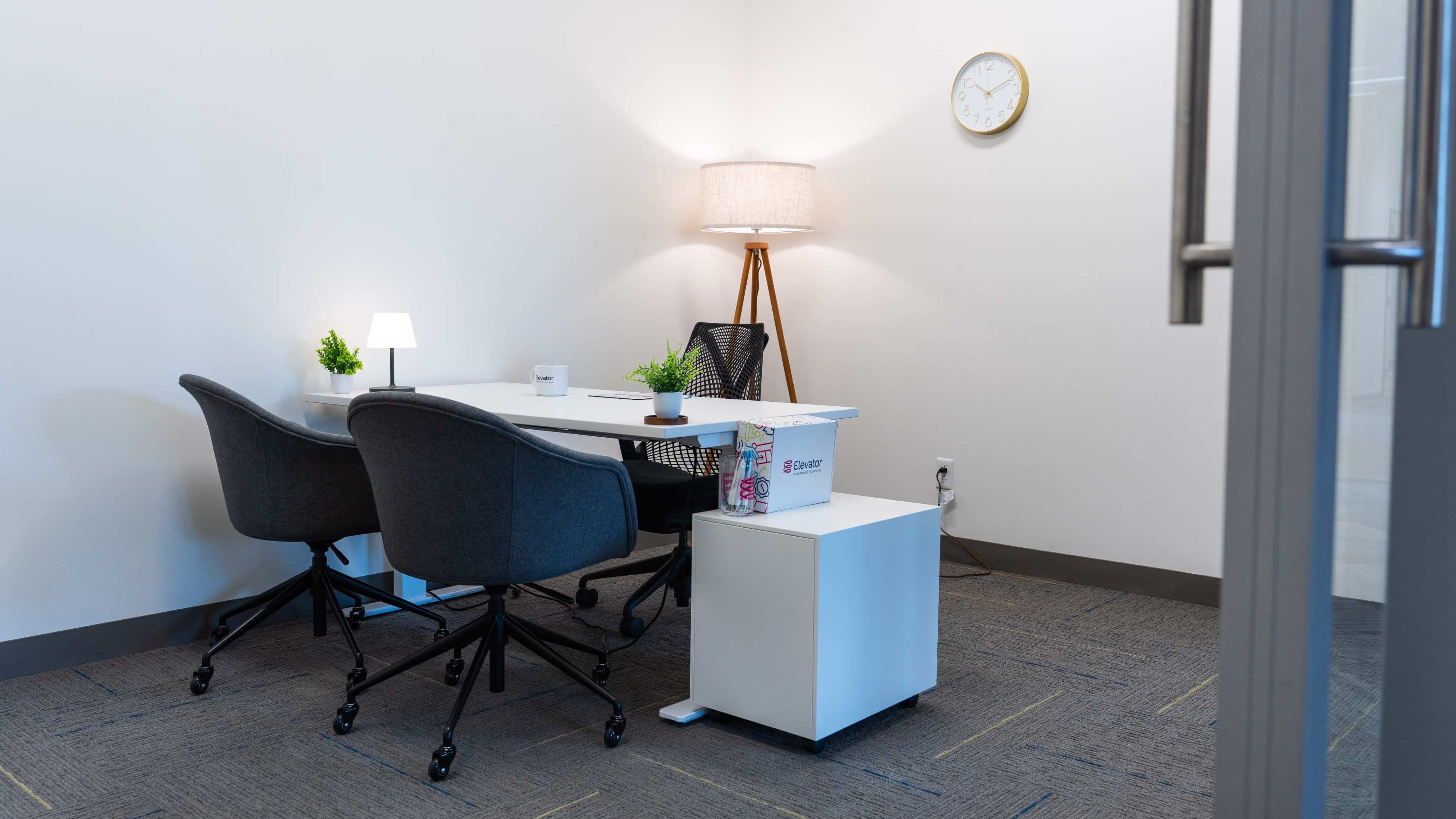 The image shows a minimalist office space with a white desk, two gray chairs, a lamp, and a small plant, all set against light-colored walls.