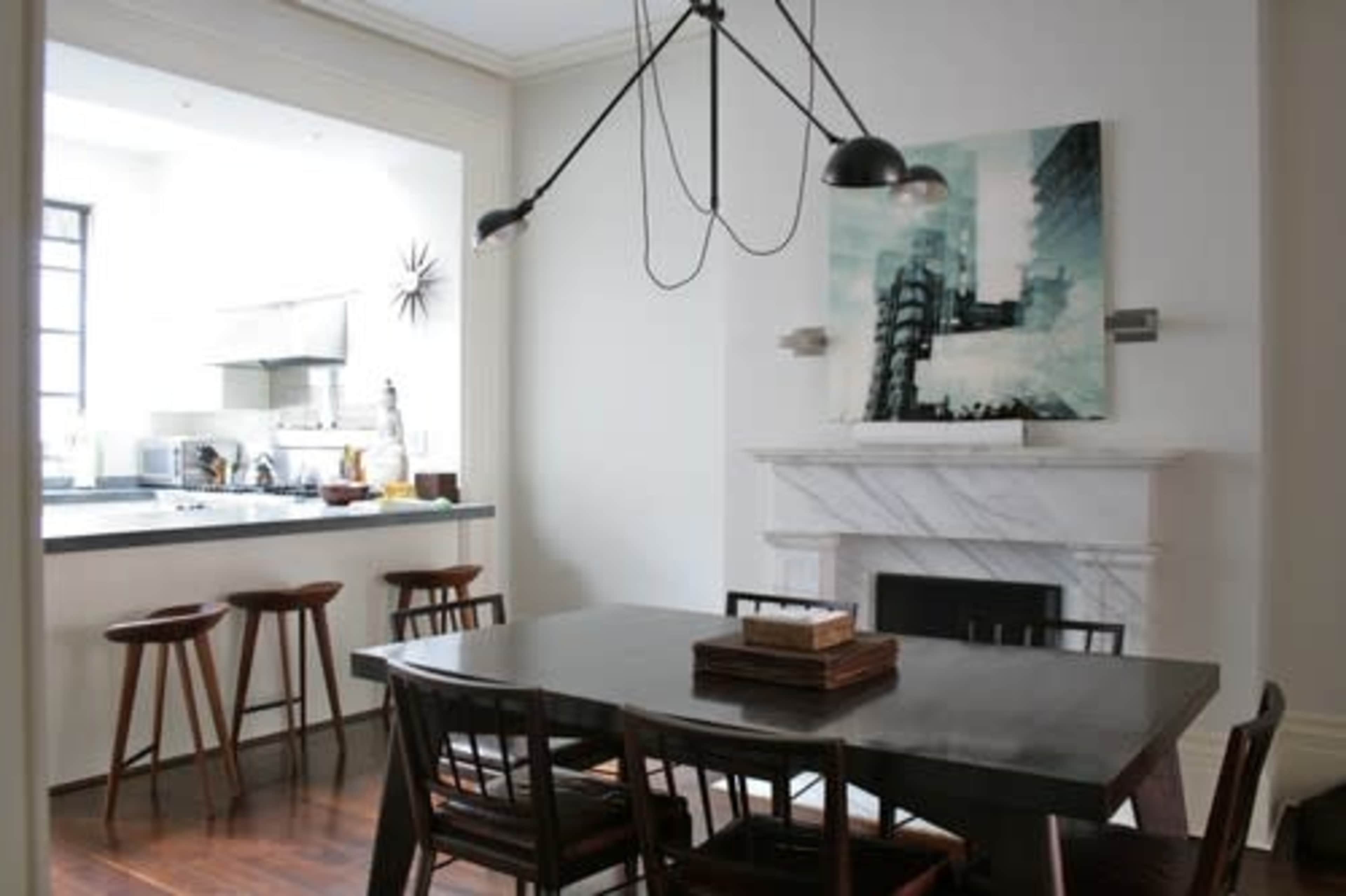 A dining area with a large wooden table surrounded by chairs, leading to an open kitchen with bar stools and a modern art piece above a marble fireplace.