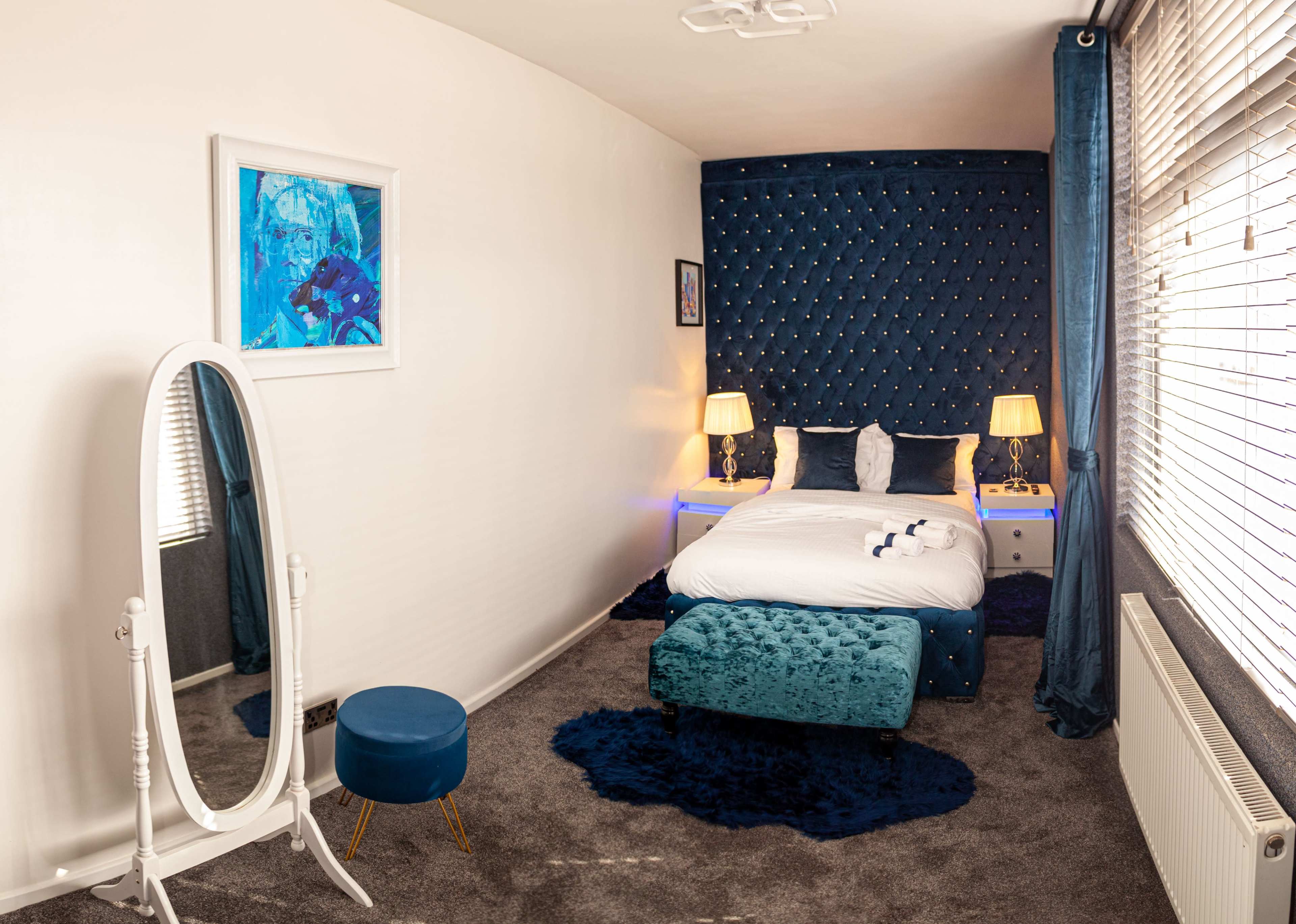 A neatly arranged bedroom featuring a bed with a tufted headboard, a mirror, and a small blue ottoman, all set against a backdrop of light-colored walls and dark blue curtains.