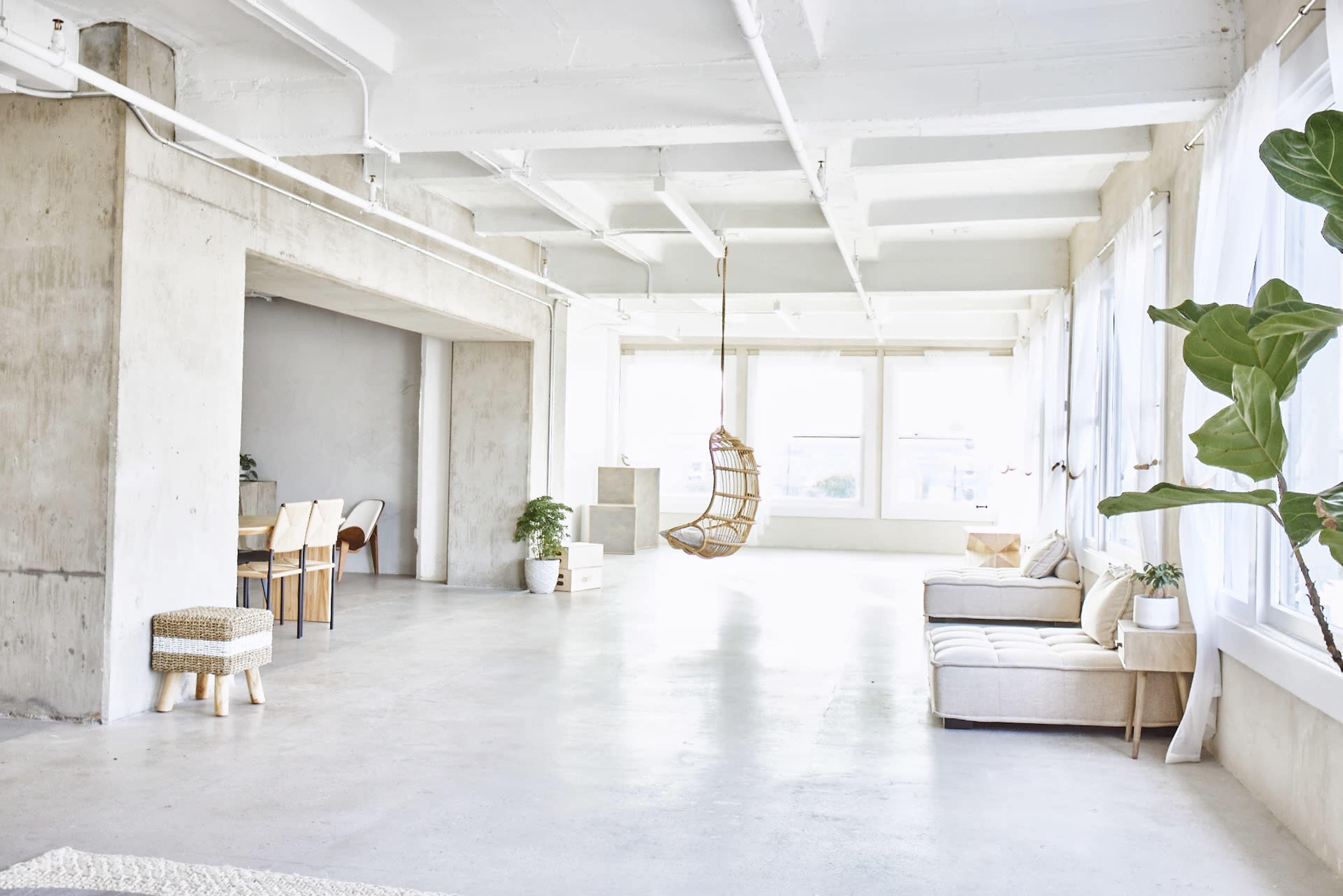 The image shows a spacious, minimalist room with concrete walls, large windows, a hanging chair, and a light-colored sofa.