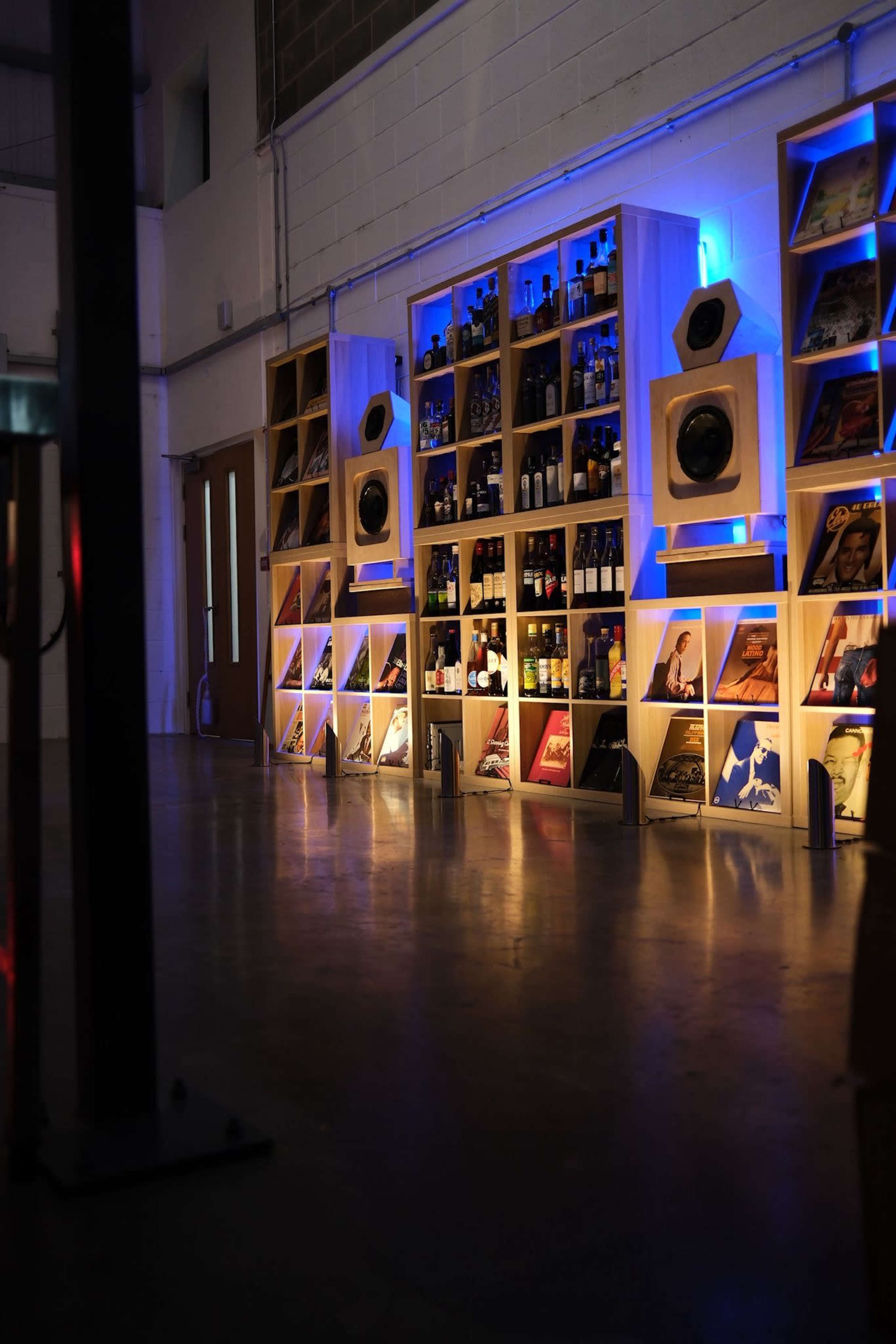 The image shows a modern interior space with a wall-mounted display featuring a variety of bottles and vinyl records, illuminated by blue lighting.