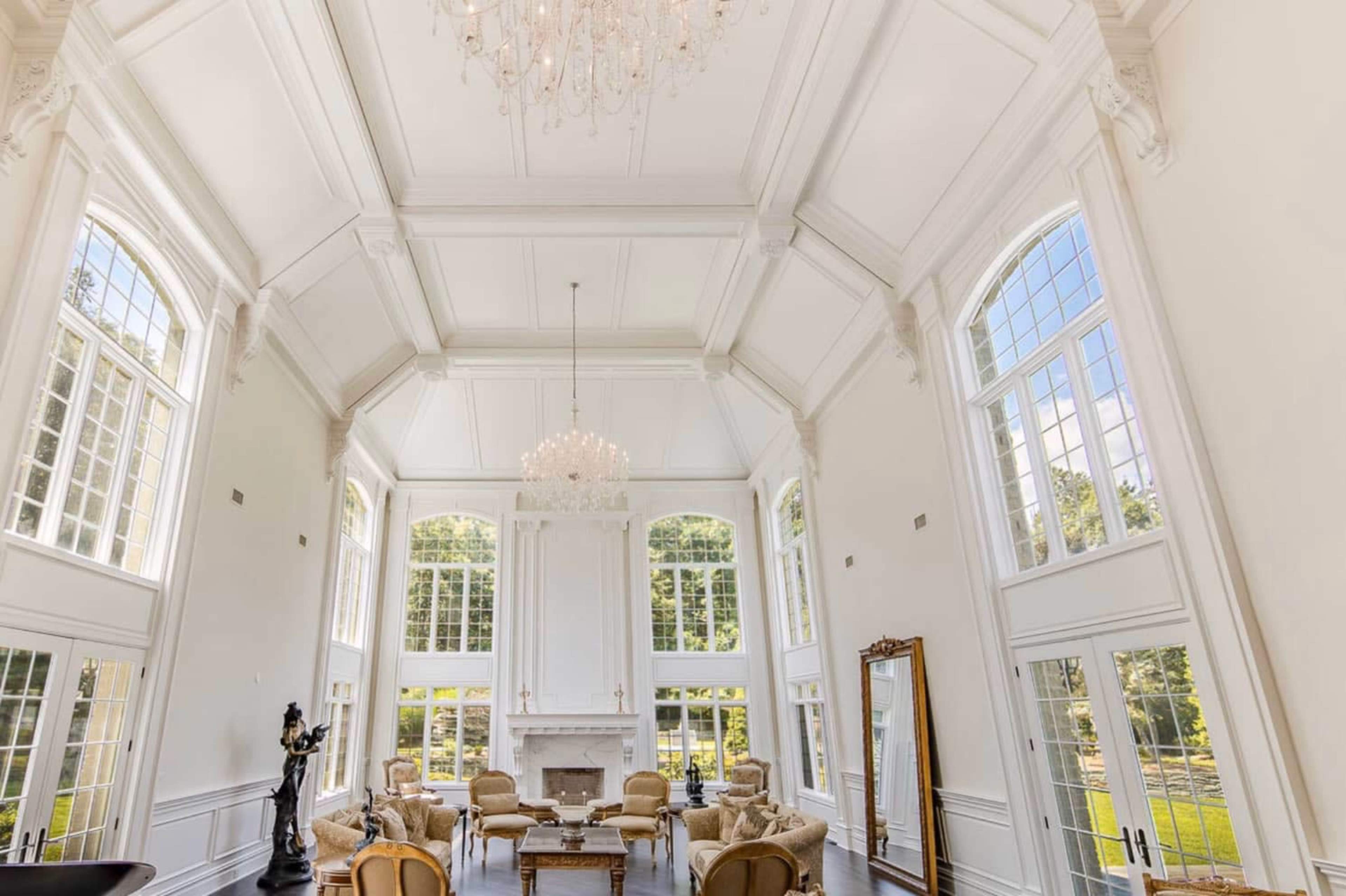 The image shows a spacious living room with high ceilings, large windows, and a chandelier, featuring elegant furniture arranged around a central fireplace.