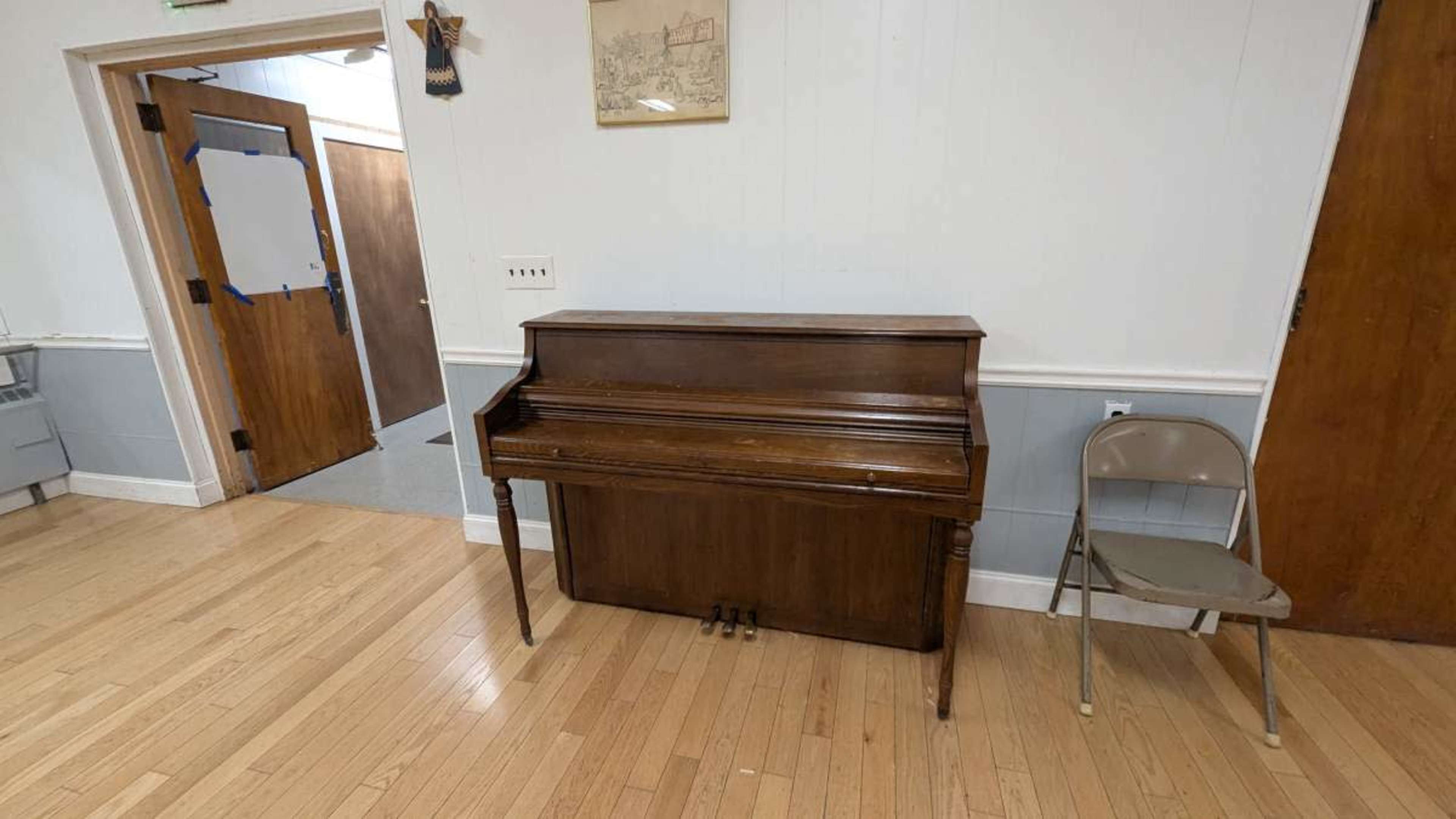 A wooden upright piano stands against a wall next to a folding chair in a room with wooden flooring and two doorways.