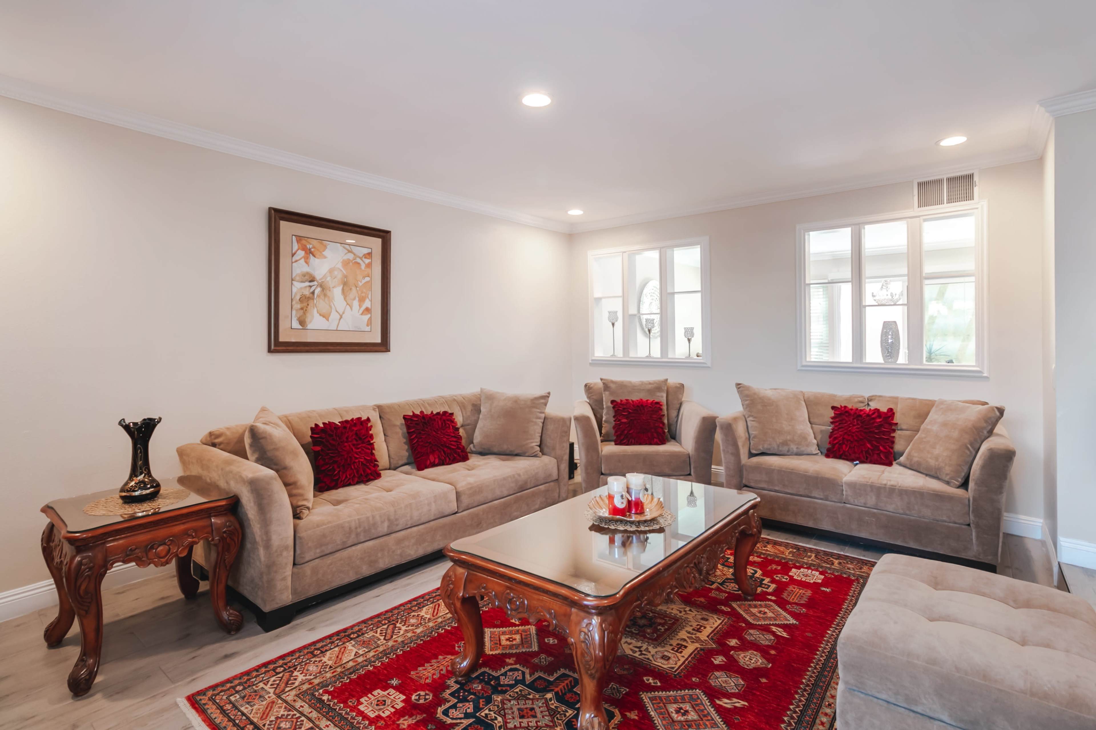 A living room features two sofas and two armchairs arranged around a coffee table on a patterned rug, with decorative pillows and a framed picture on the wall.
