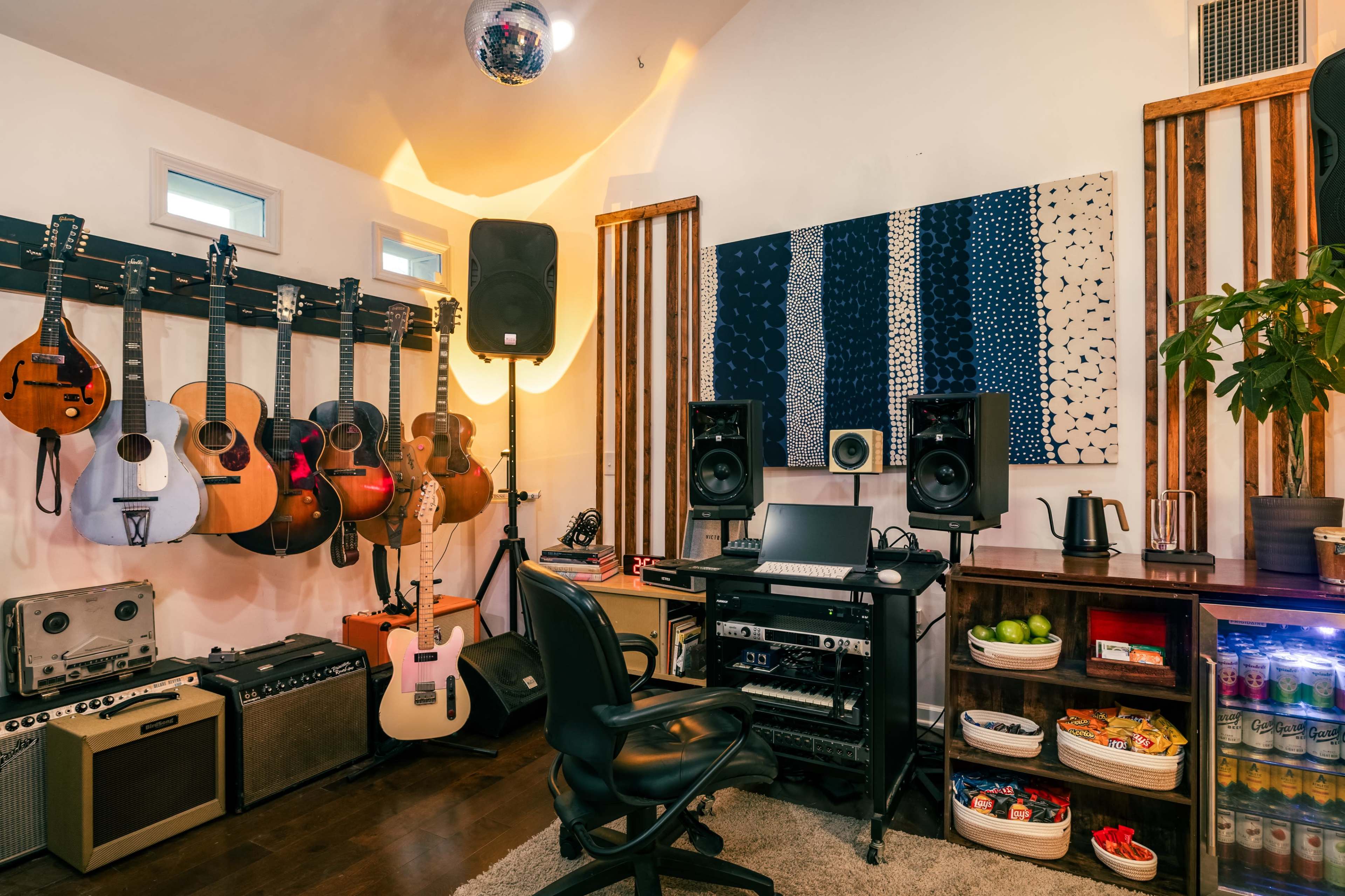 The image shows a home recording studio featuring various guitars hanging on a wall, amplifiers, a desk with audio equipment, and a decorative wall art piece.