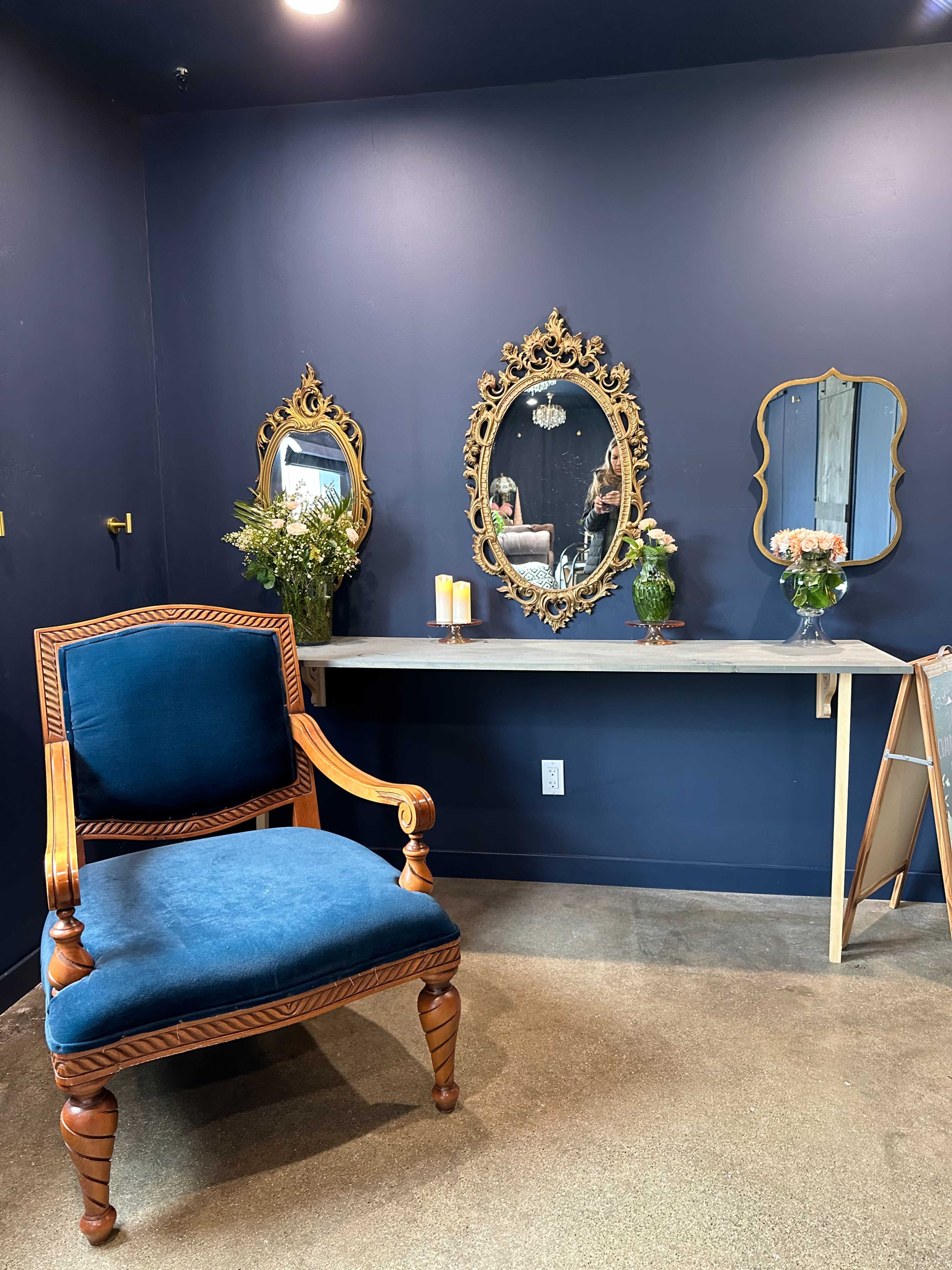 The room features a blue wall, a wooden table with vases of flowers, two ornate mirrors, and a blue chair with a decorative frame.