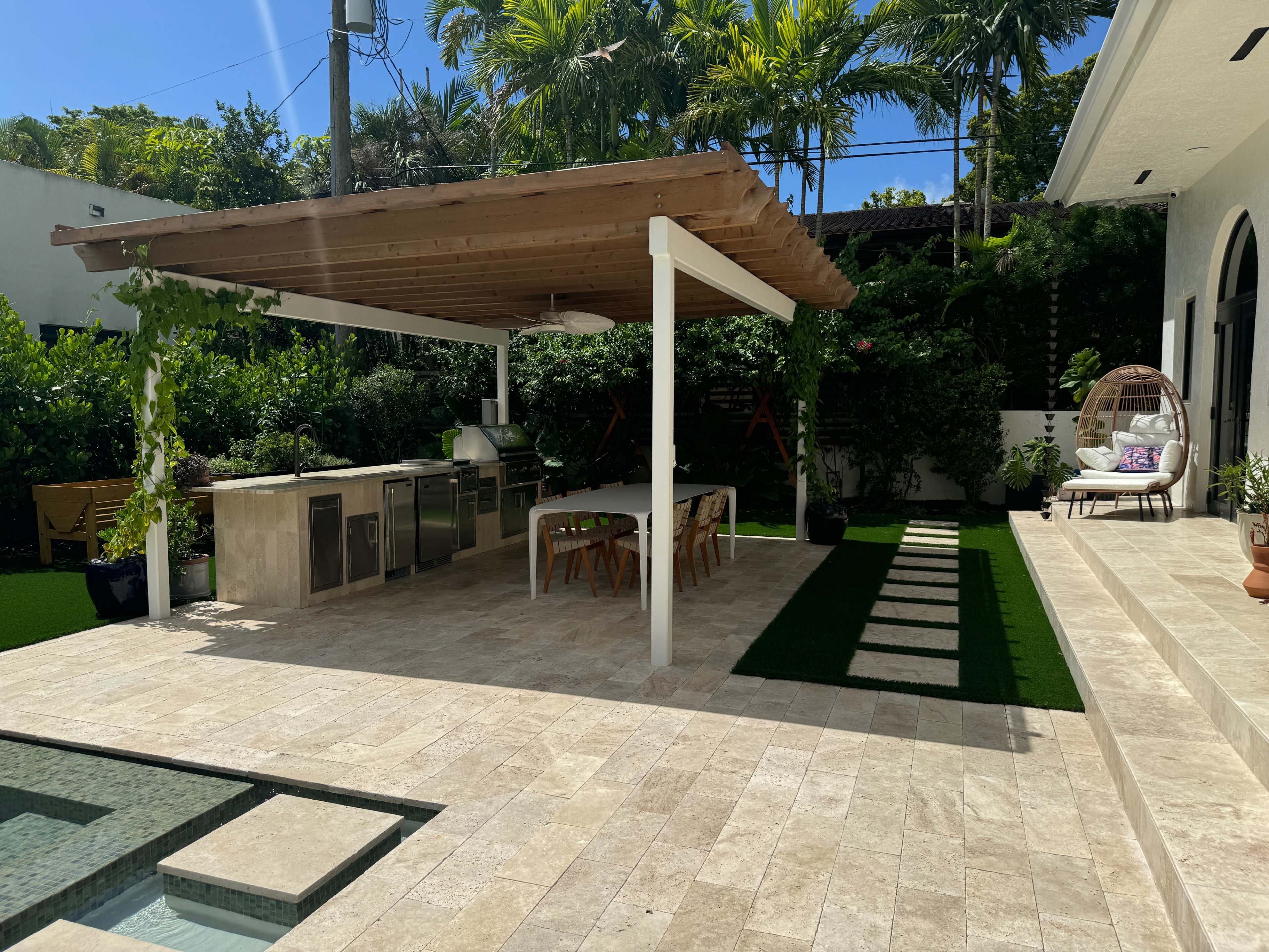 The image shows a backyard with a covered outdoor kitchen and dining area, surrounded by greenery and a small pool.
