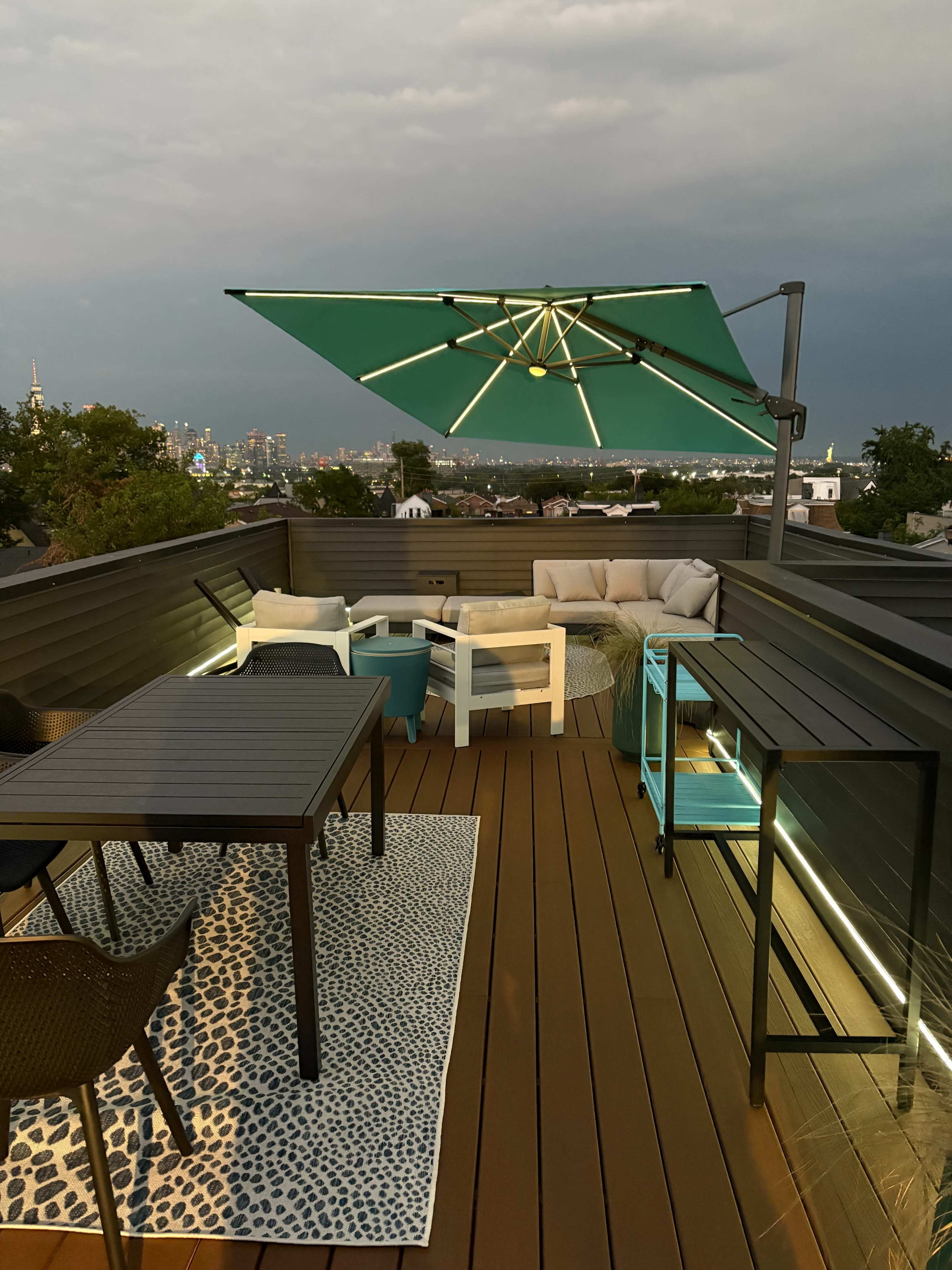 A rooftop terrace features modern outdoor furniture, including a table, chairs, and a shaded lounge area, with a city skyline visible in the background.