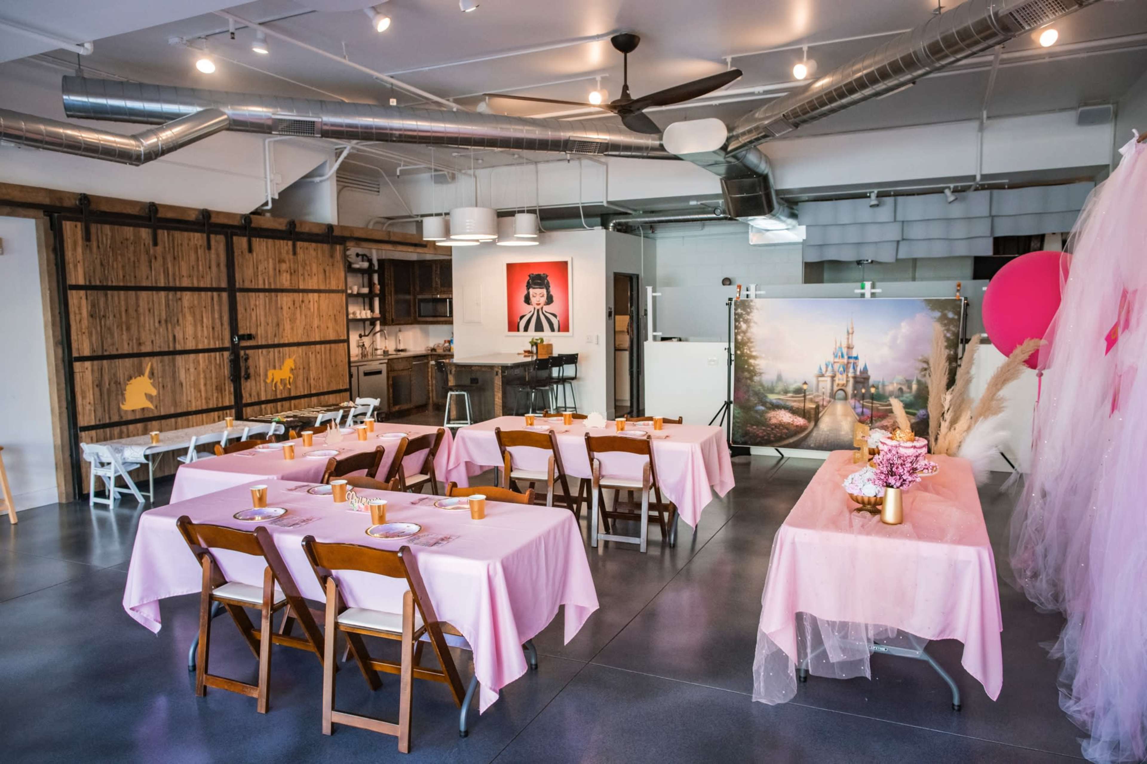 The image shows a decorated indoor space with pink tables set for an event, featuring wooden chairs and a backdrop of a castle painting.